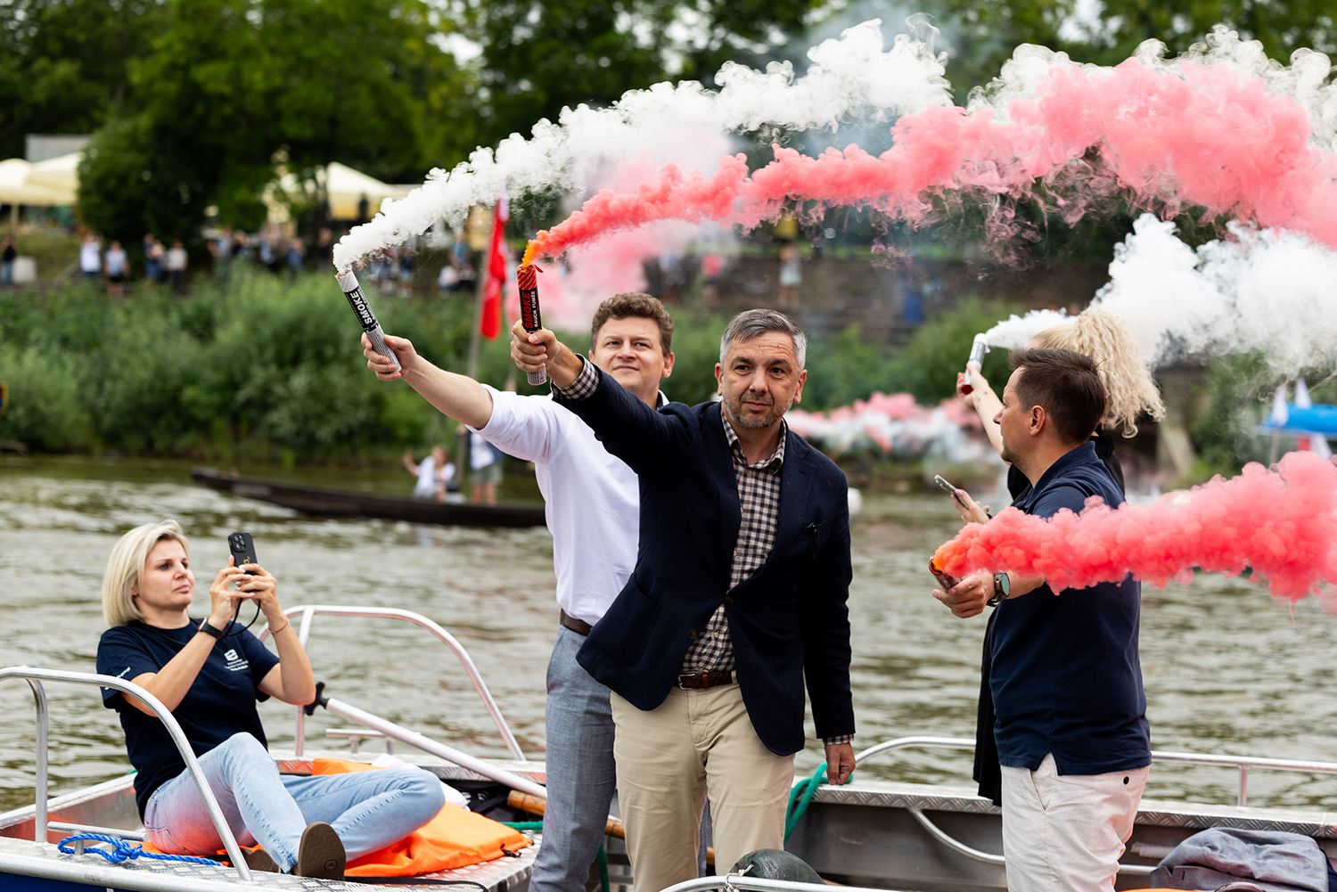 Group on a boat waving red and white smoke signals during an outdoor river event.