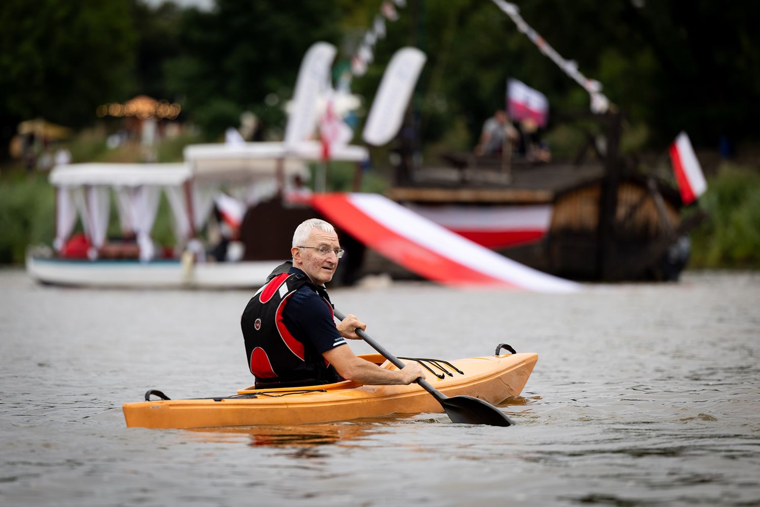 Bald man paddling wooden kayak with Polish flag boats in background.