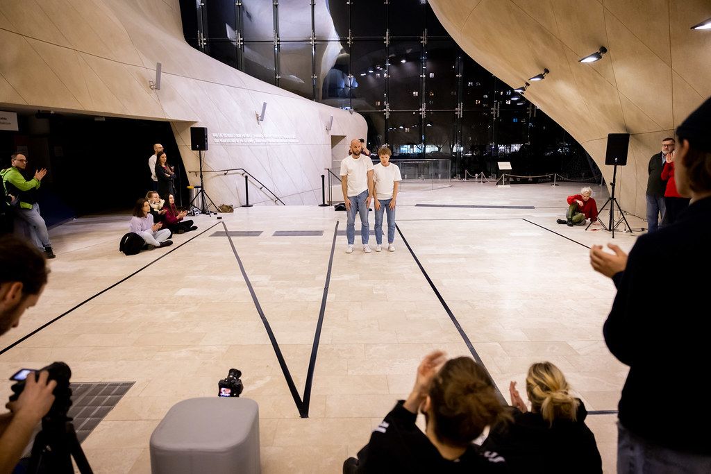 Two performers in white shirts stand together in a spacious modern venue with audience around.