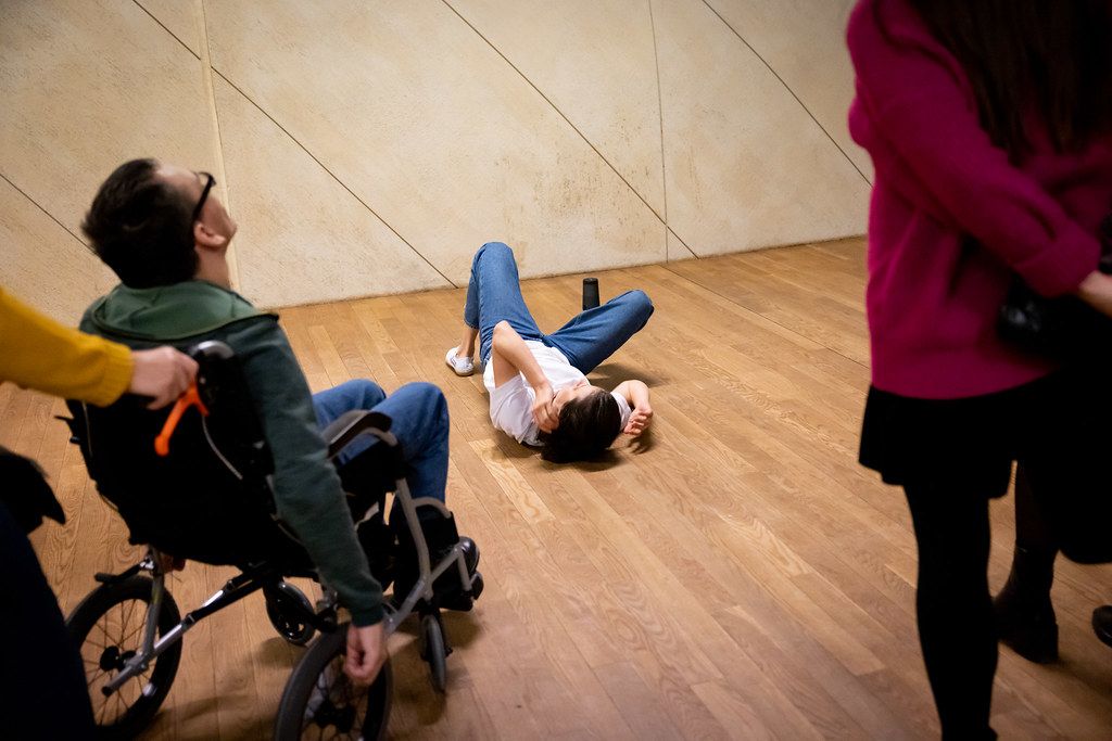 Person lying on wooden floor near wheelchair during interactive art performance.