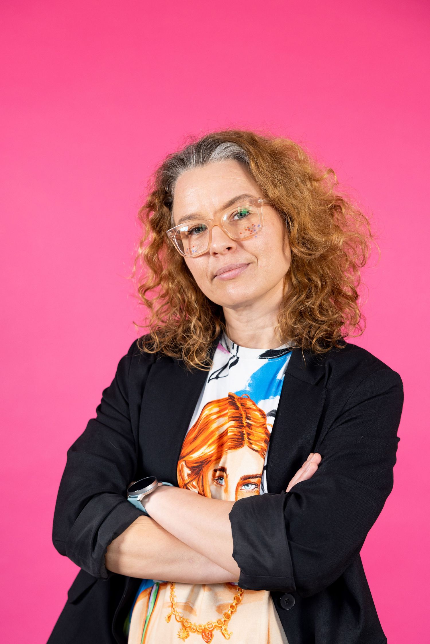 Curly red-haired woman in black blazer posing confidently against a bright pink background.