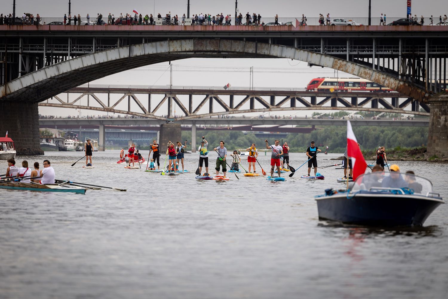 Stand-up paddleboarders and boats near a bridge with a Polish flag during a river event.