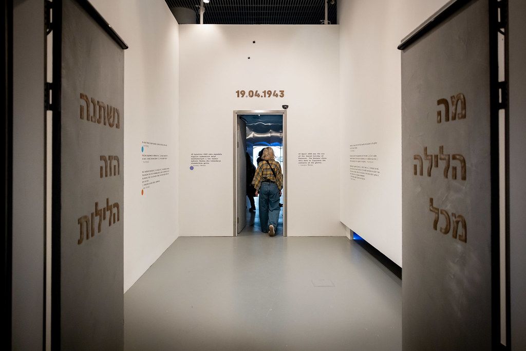 Person photographing Hebrew text installation through narrow doorway in museum.