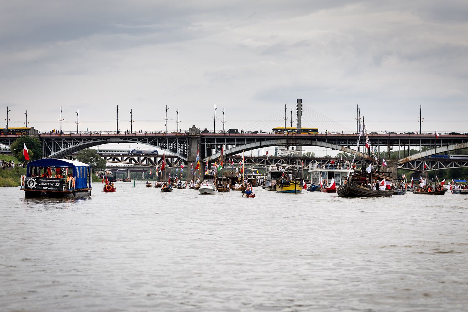 Large flotilla of boats and vessels gathered near bridge on river.