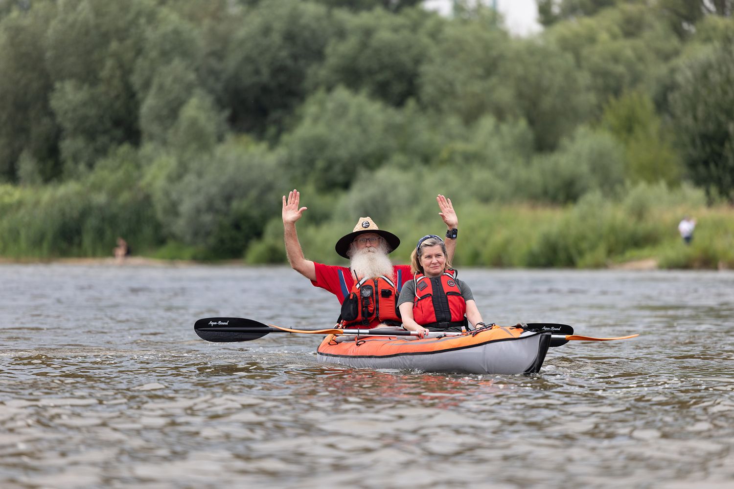 Two kayakers in red life jackets waving hands on sunny river with green banks.