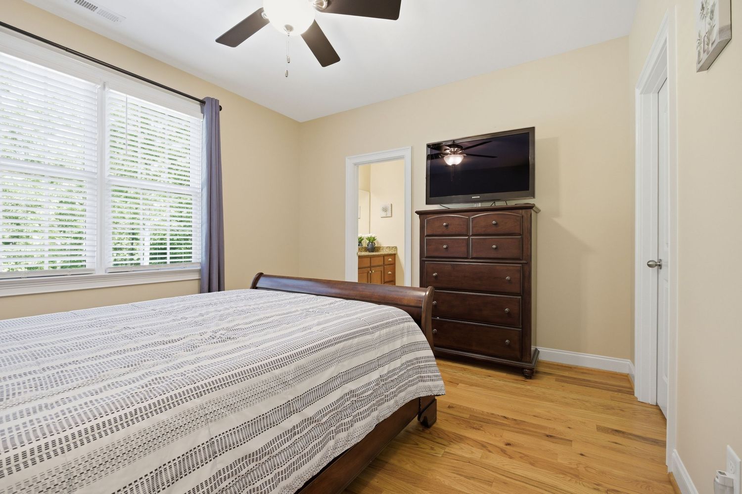Bedroom with hardwood floors, dark wood dresser, wall-mounted TV, ceiling fan, and natural light.