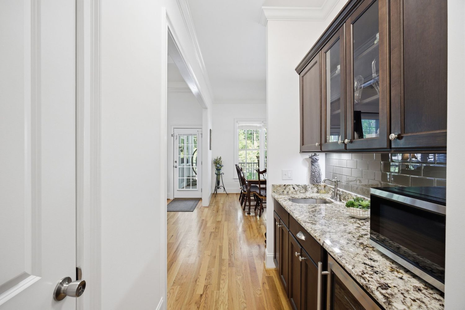 Galley kitchen with dark wood cabinets, granite countertops, glass-front uppers, and hardwood floors leading to a bright dining area.