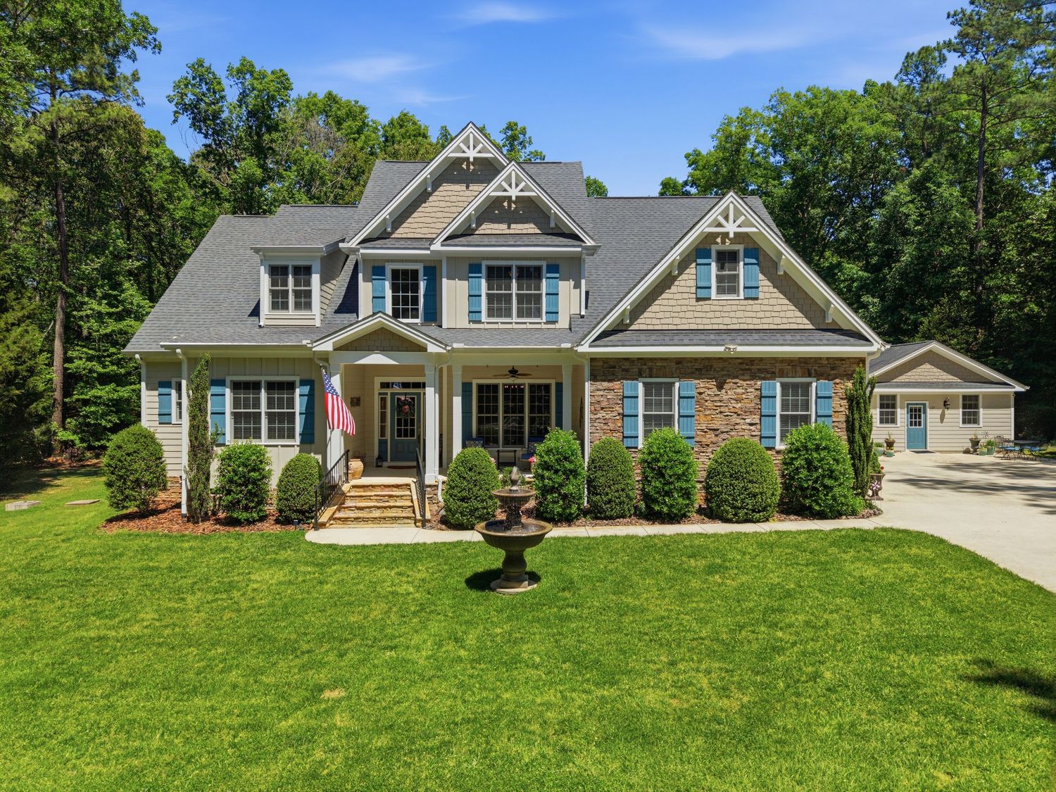 Grand craftsman-style home with stone detailing, blue shutters, center fountain, and green lawn.