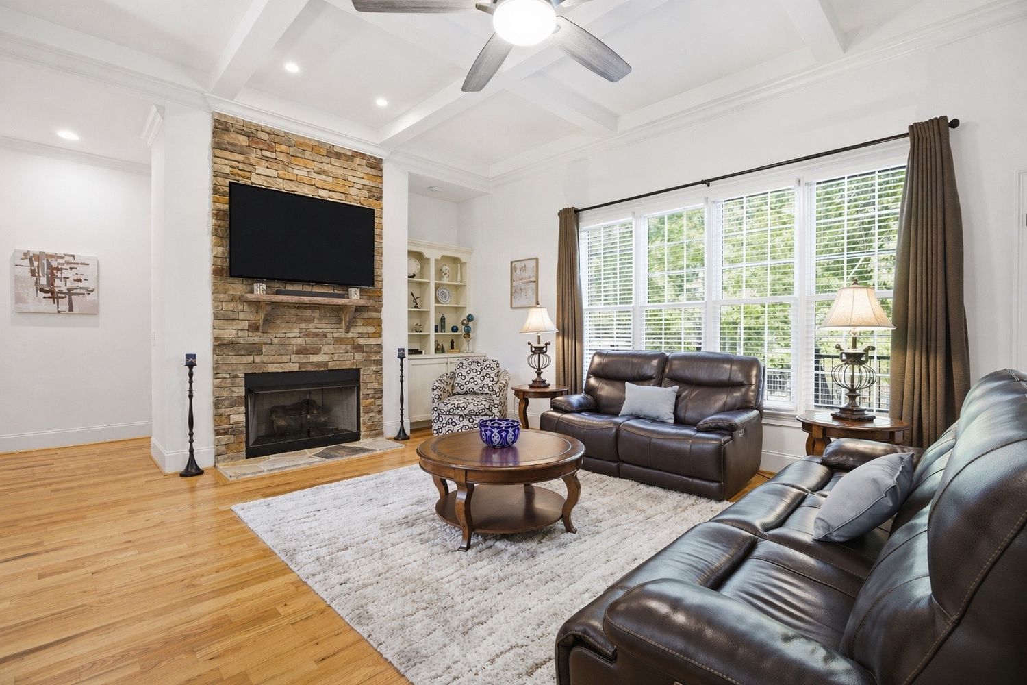 Spacious living room featuring coffered ceiling, stone fireplace, leather furniture, and bright windows.