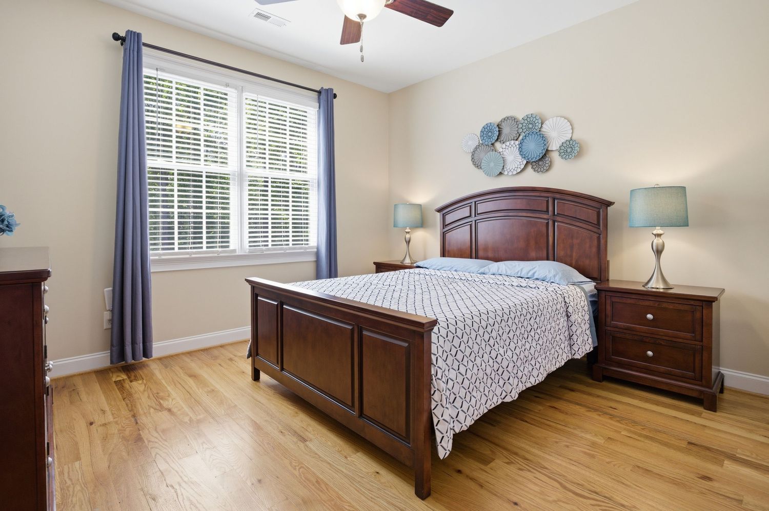 Bright guest bedroom with hardwood floors, ceiling fan, dark wood furniture, and plantation shuttered window.