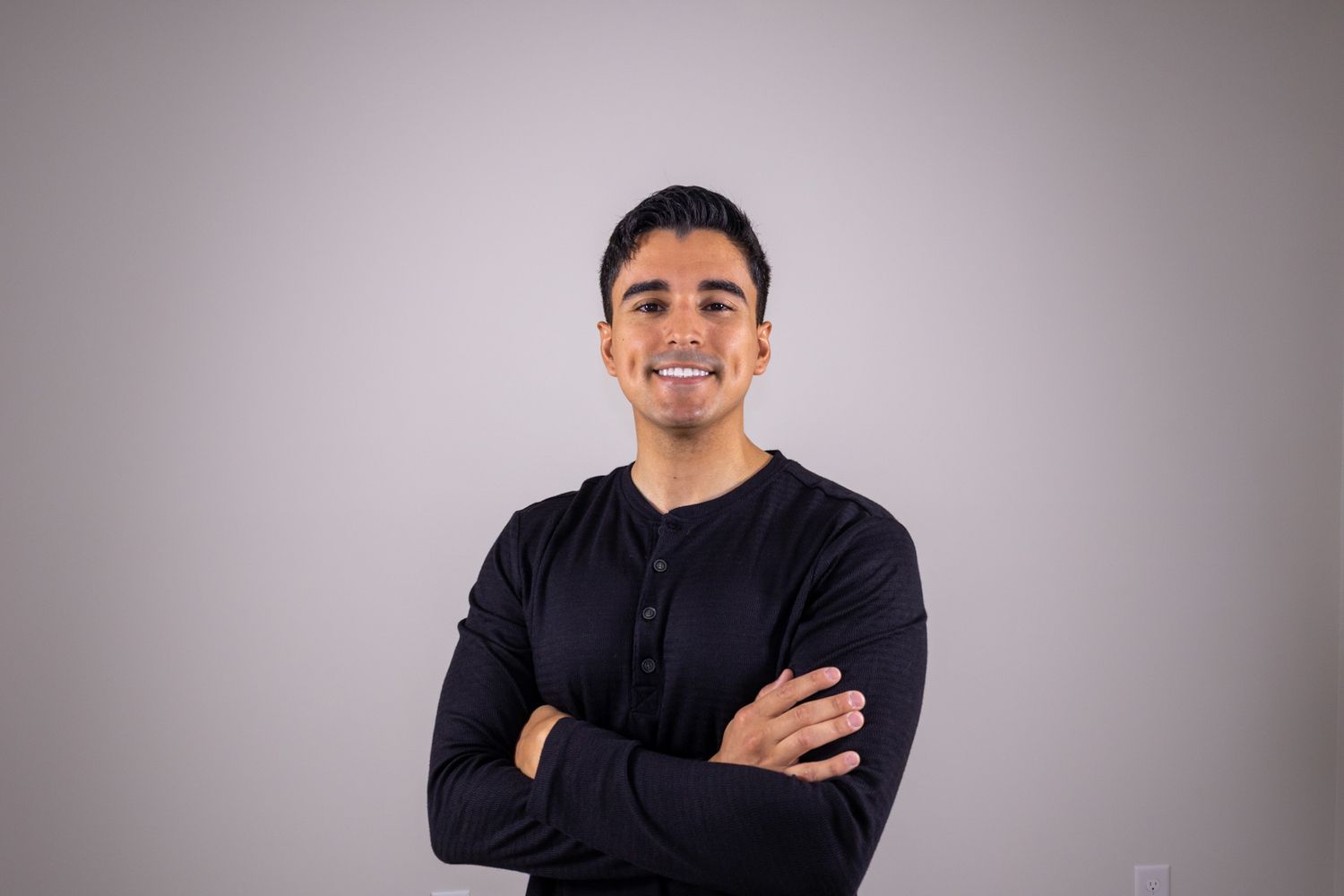 Smiling young man in black long-sleeve shirt standing with arms crossed on gray background.