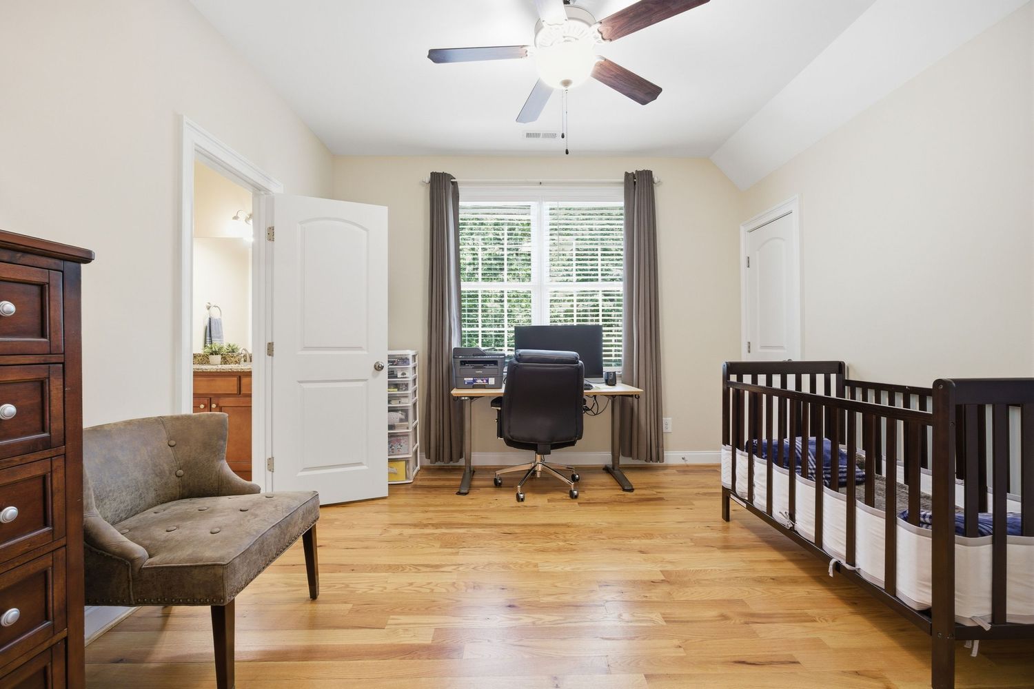Versatile bedroom with hardwood floors, dark wood crib, office desk, ceiling fan, and natural light.
