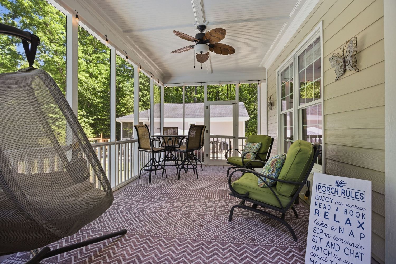 Screened porch with ceiling fan, wicker hanging chair, and outdoor seating overlooking trees.