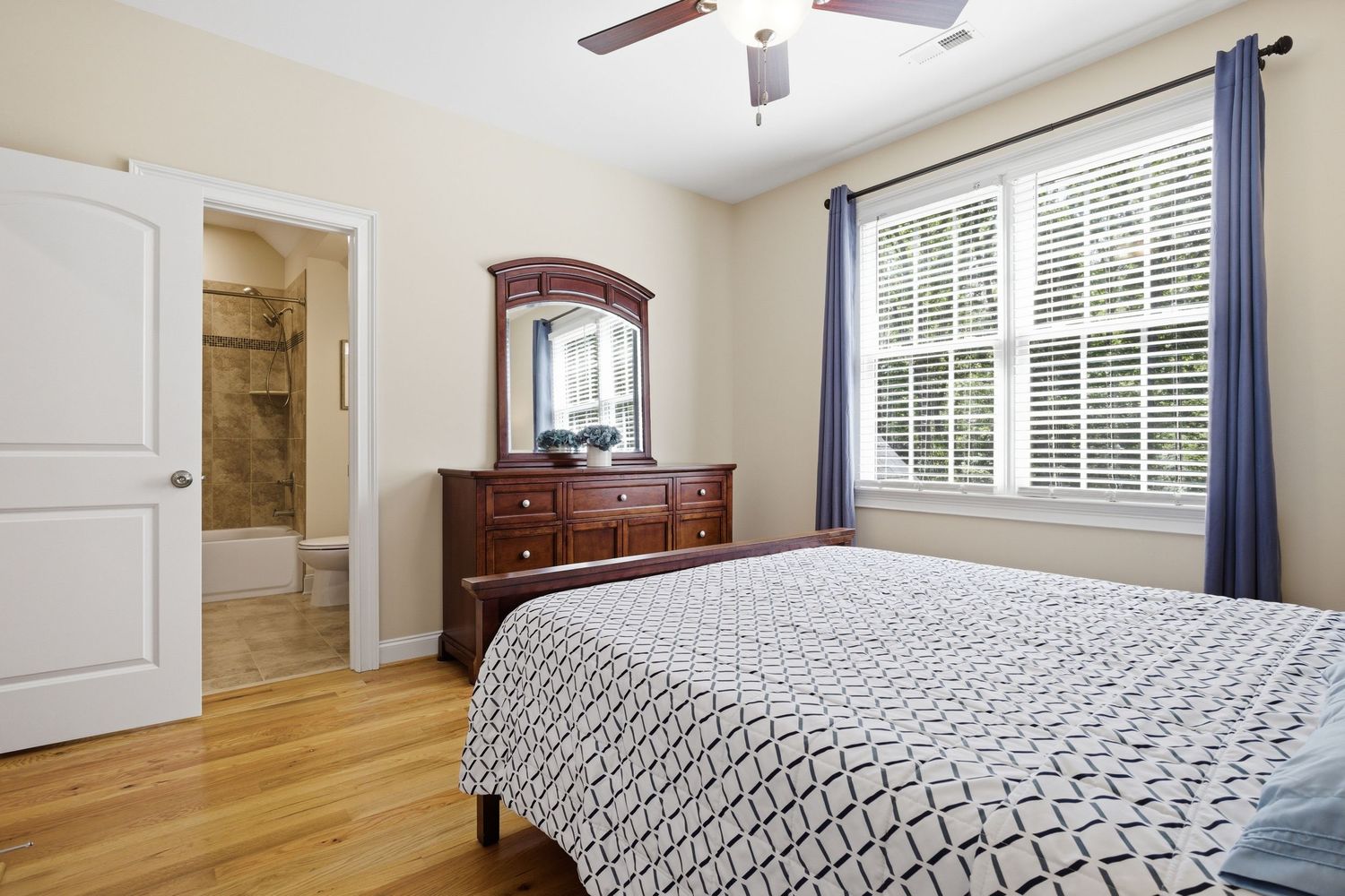 Comfortable bedroom with hardwood floors, ceiling fan, dark wood dresser, and en-suite bathroom access.
