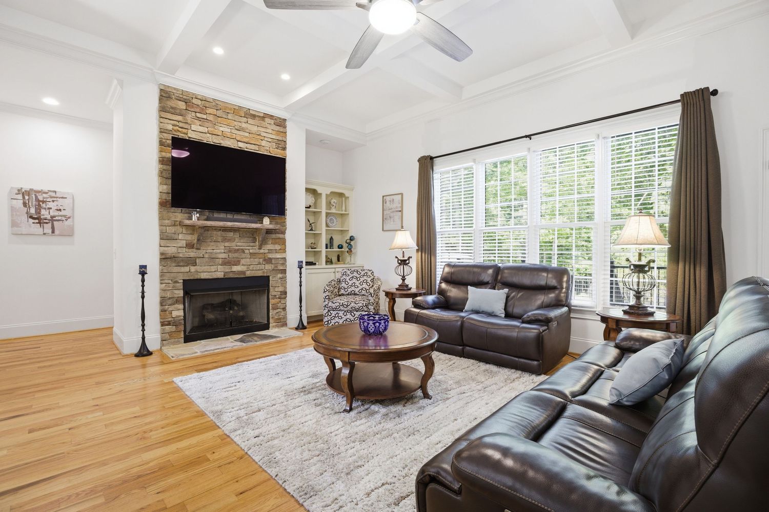 Living room with coffered ceiling, stone fireplace, leather sofas, and large windows with natural light.