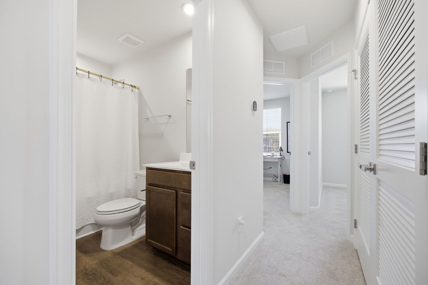 Clean white bathroom hallway with dark vanity, toilet, shower curtain, and louvered closet doors.