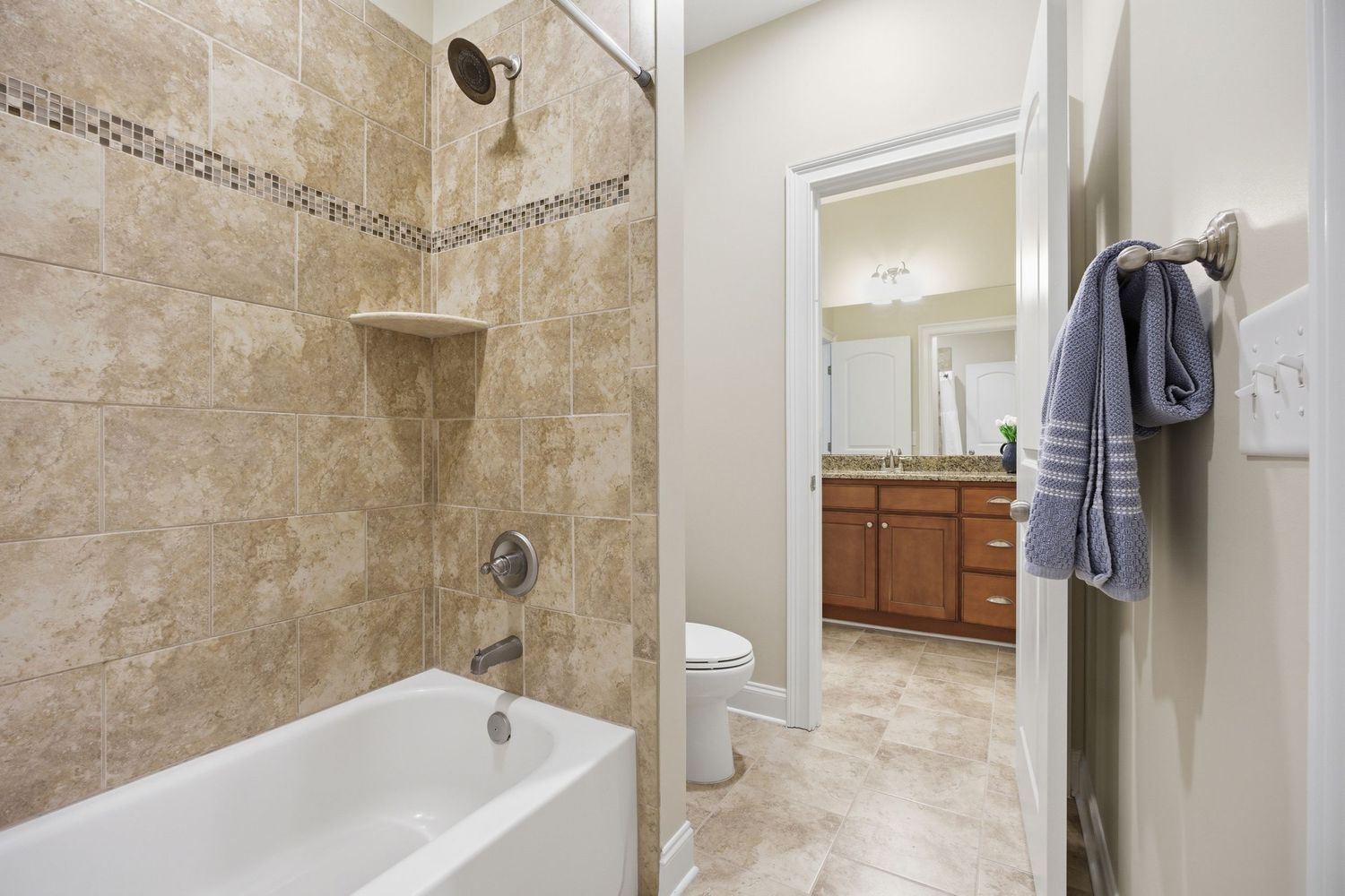 Tiled shower-tub combo with decorative border, white towels, toilet, and wood vanity visible beyond.