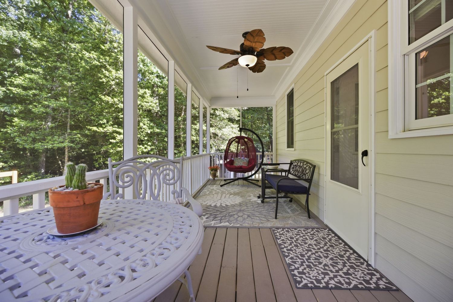 Screened porch with ceiling fan, hanging egg chair, patio dining set, and wooded views on a sunny day.