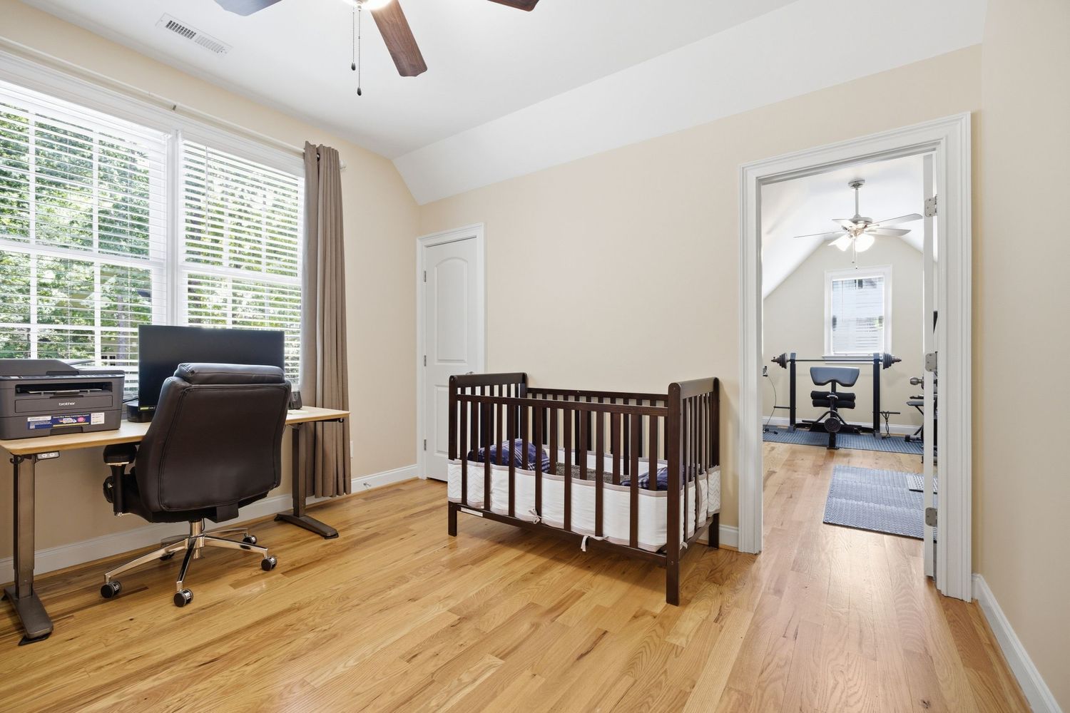 Bright bedroom with hardwood floors, dark wood crib, ceiling fan, and view into adjacent room.