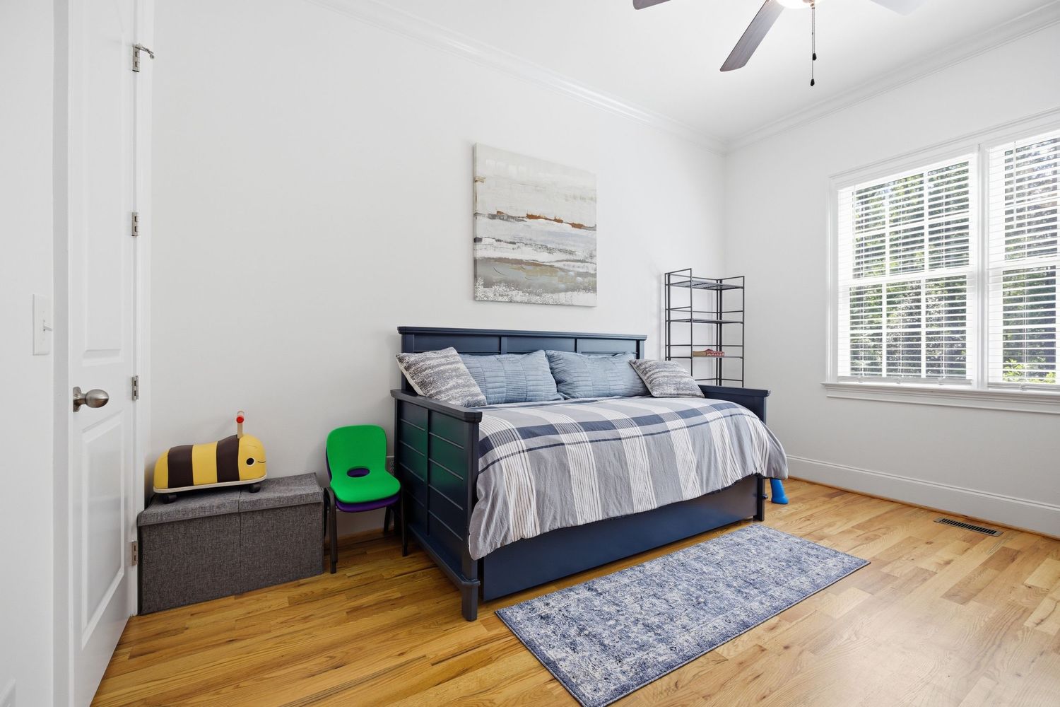 Bright bedroom with navy bed, hardwood floors, ceiling fan, area rug, and white walls with natural light.