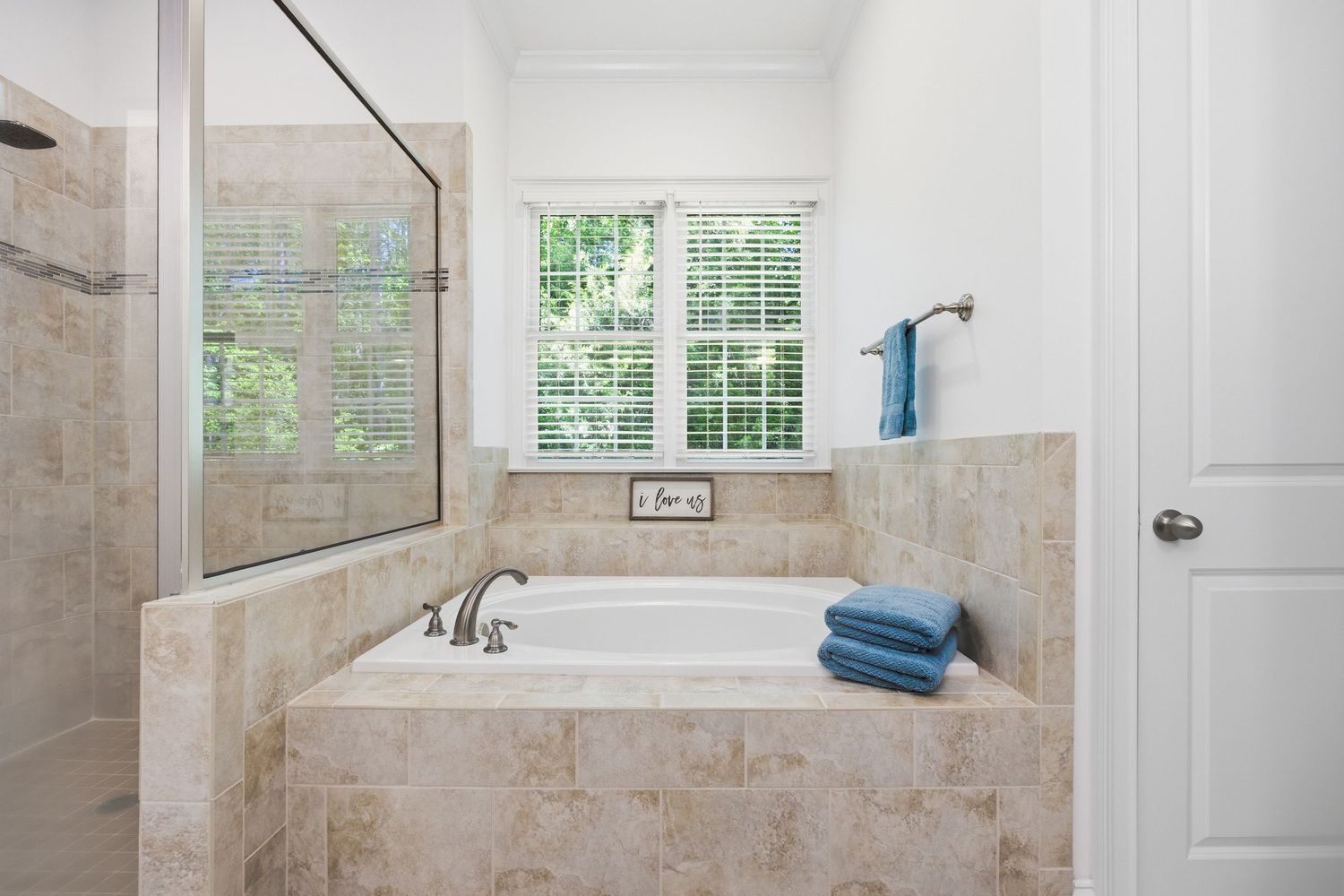 Tiled soaking tub with garden window view surrounded by beige stone tile in elegant master bathroom.
