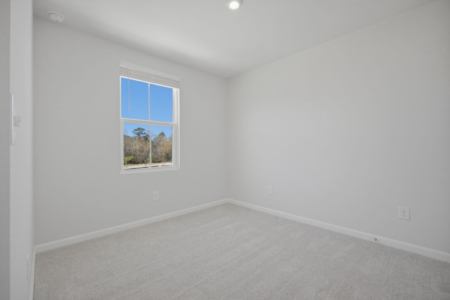 Bright empty bedroom with light carpet, white walls, single window, and recessed lighting.