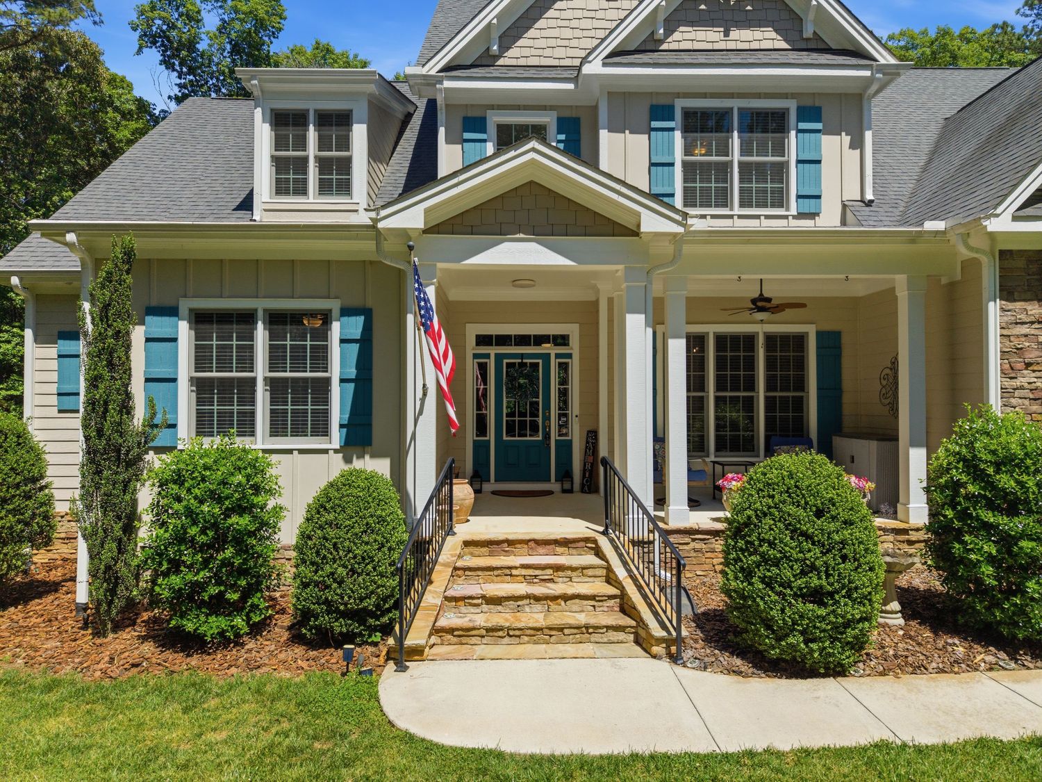 Close-up of craftsman home entrance with blue door, stone steps, American flag, and manicured shrubs.