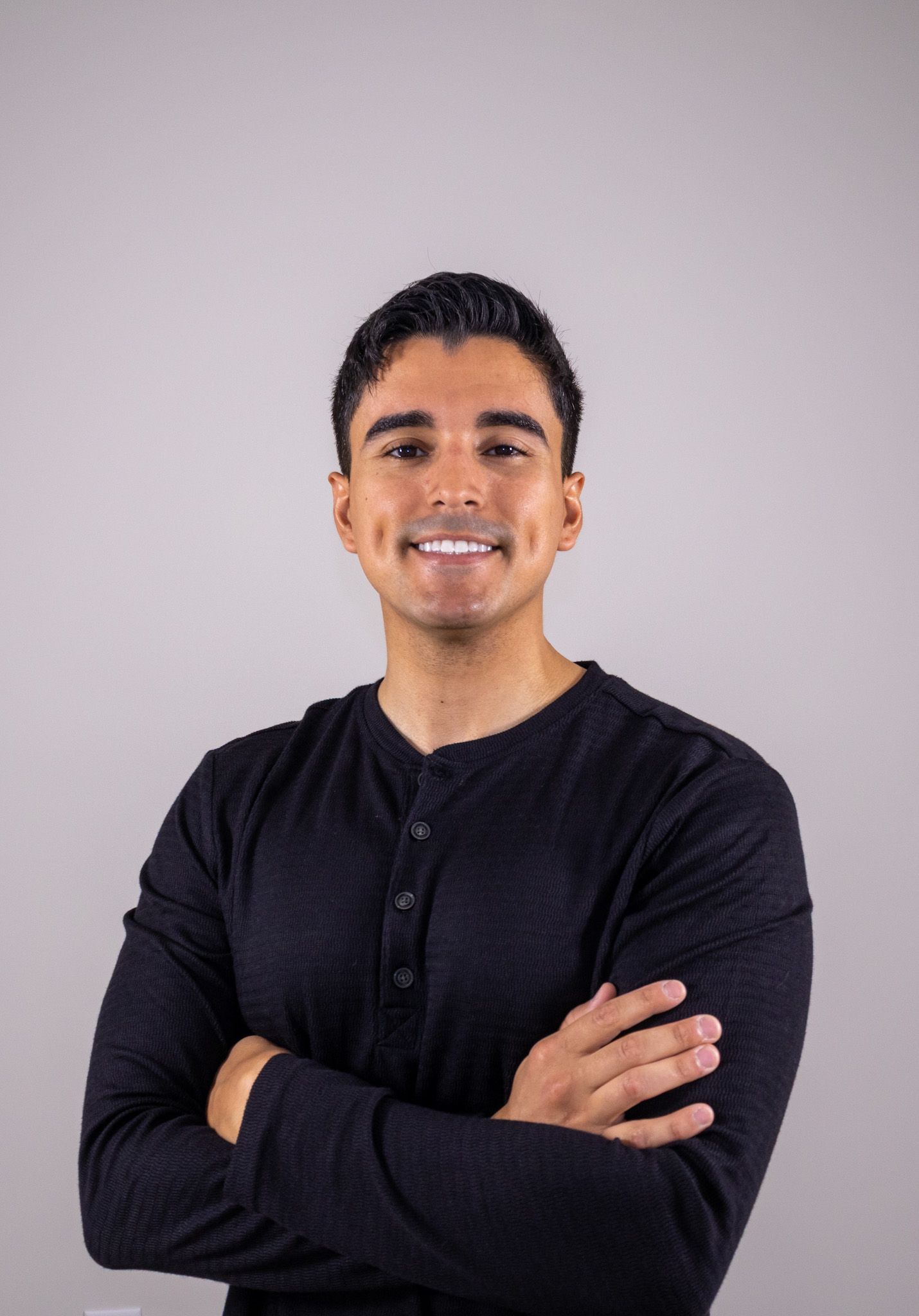 Young man in black henley shirt smiling with arms crossed against gray background.