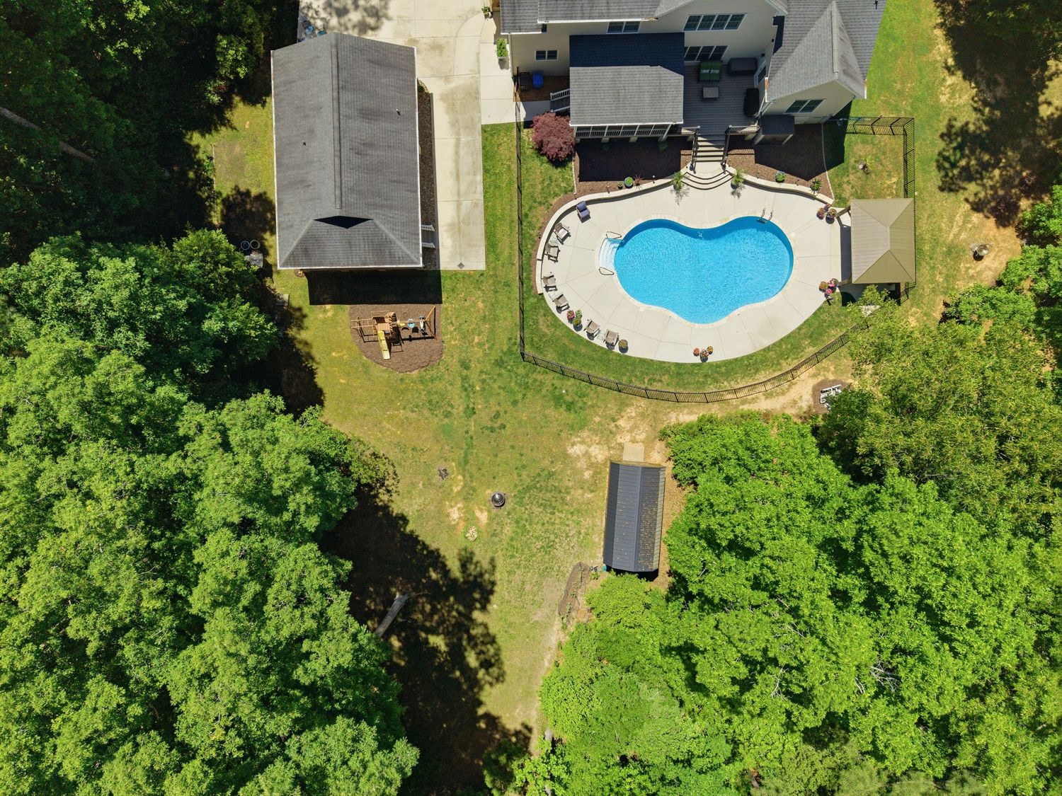 Aerial view of backyard with kidney-shaped pool, patio, and lush green tree surroundings.