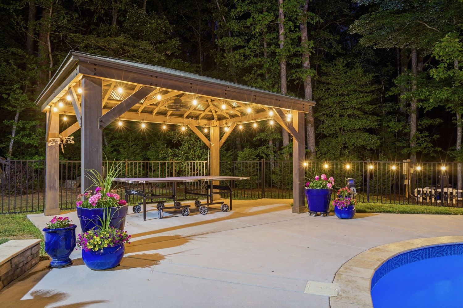 Beautiful outdoor pavilion with string lights, dining area, and pool adjacent to lush trees.