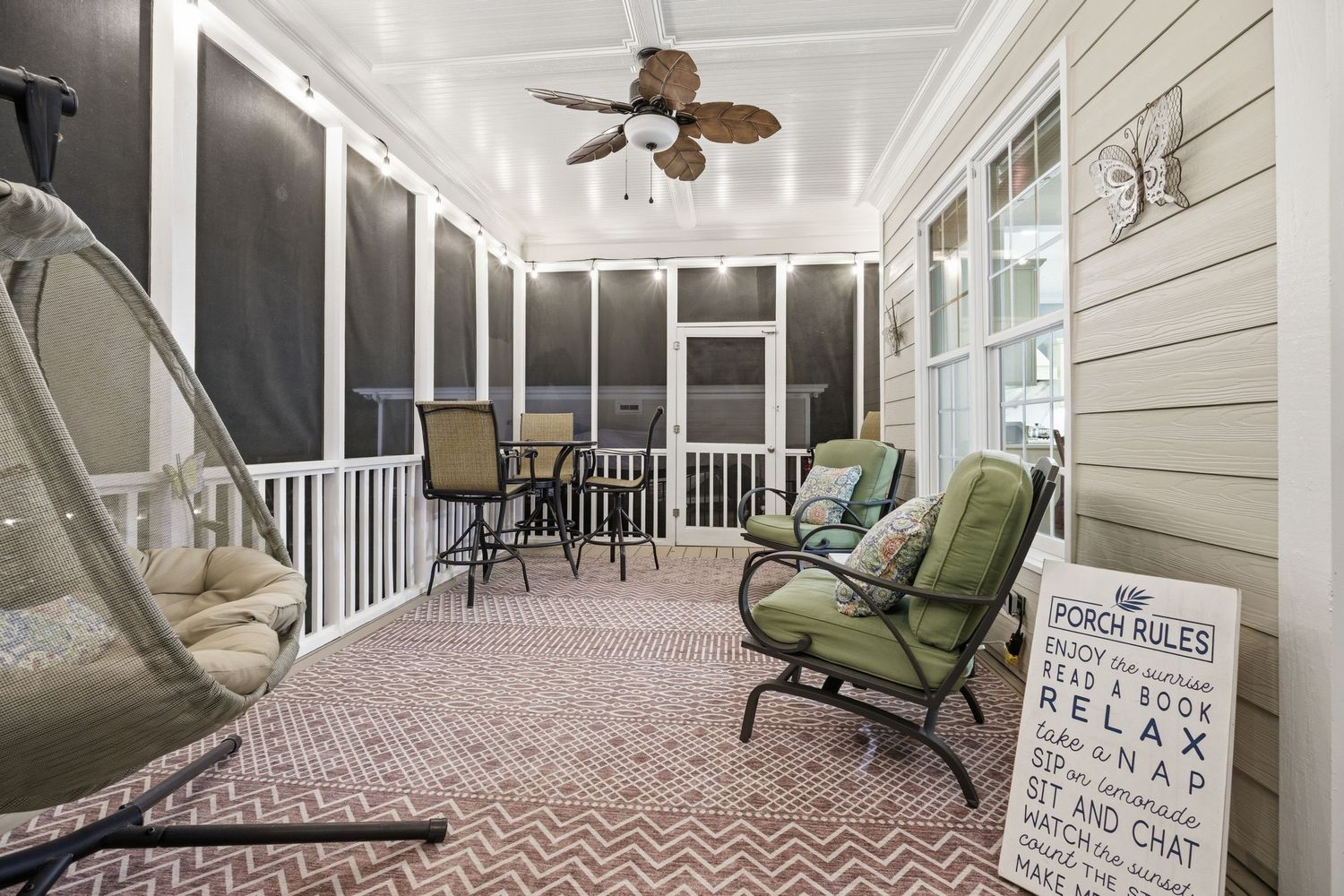 Charming screened porch with ceiling fan, hanging chair, green seating, and decorative sign.