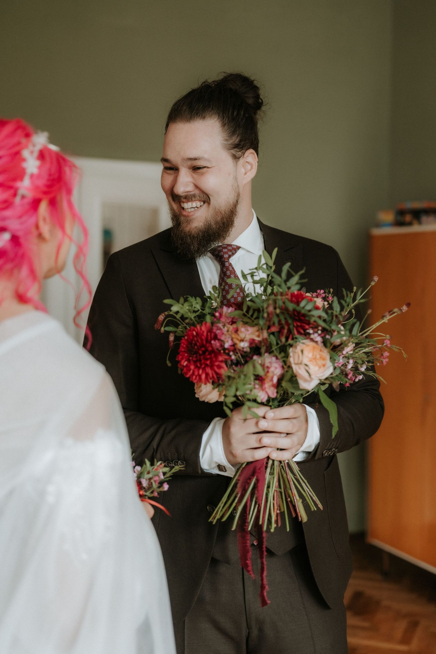 Smiling groom in dark suit holds colorful bridal bouquet of red dahlias and pink roses for bride with pink hair.