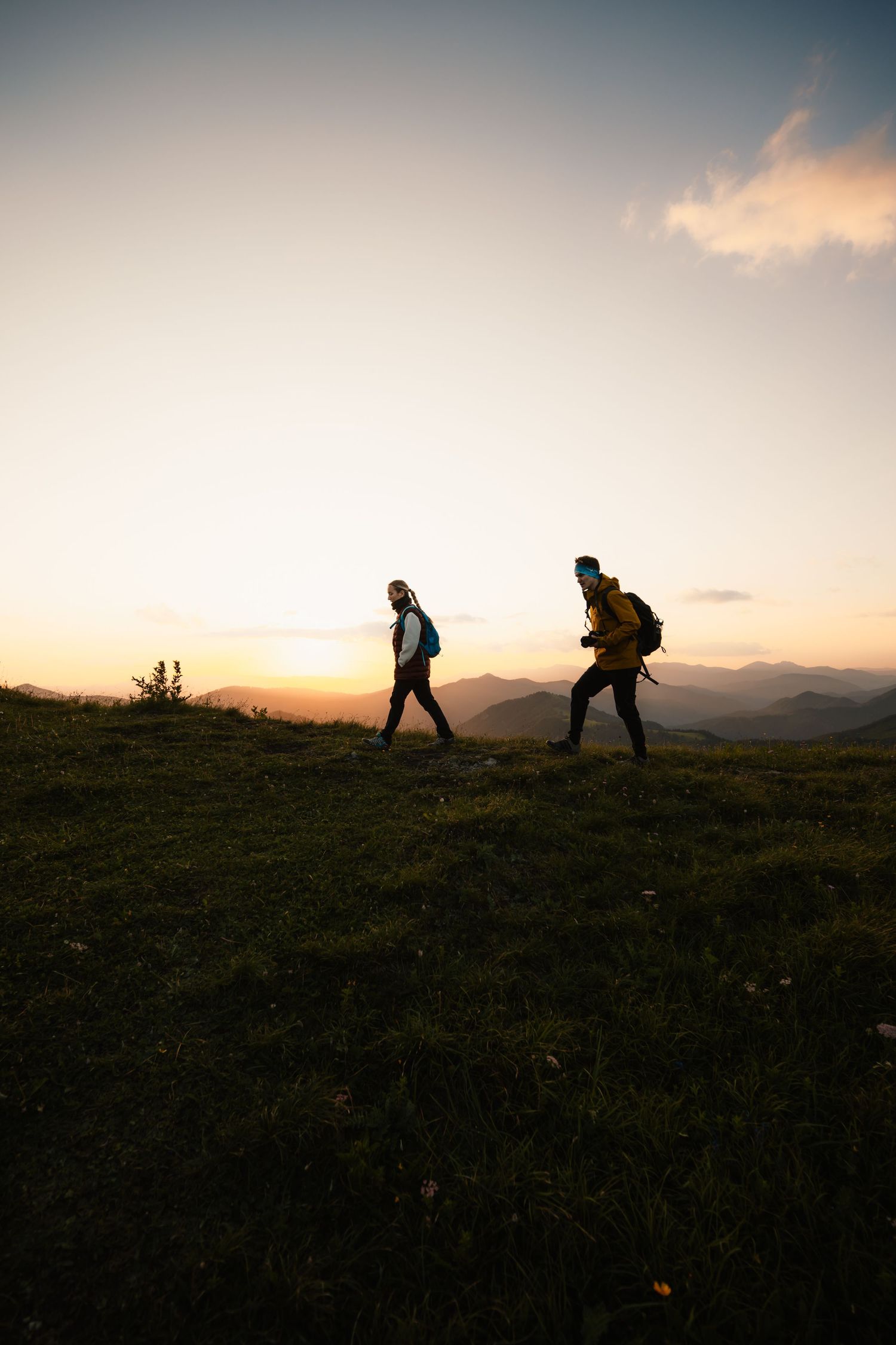 Two hikers with backpacks silhouetted against a vibrant mountain sunset on a grassy ridge trail.