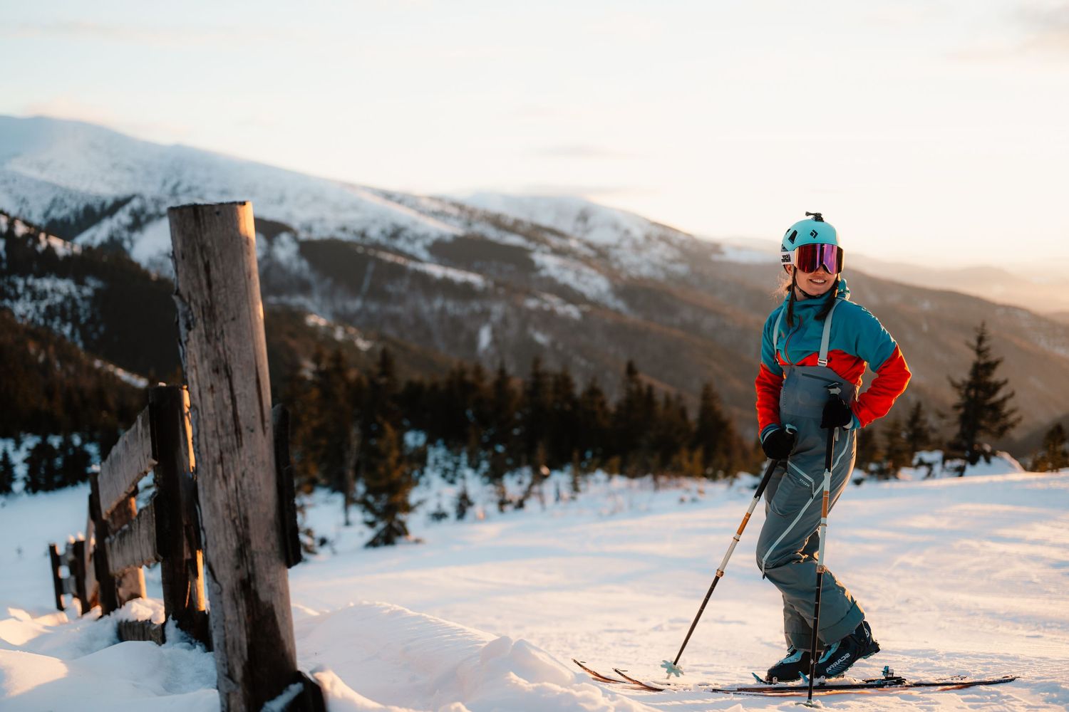 Female skier in colorful gear standing on snowy mountain slope at golden hour sunset.