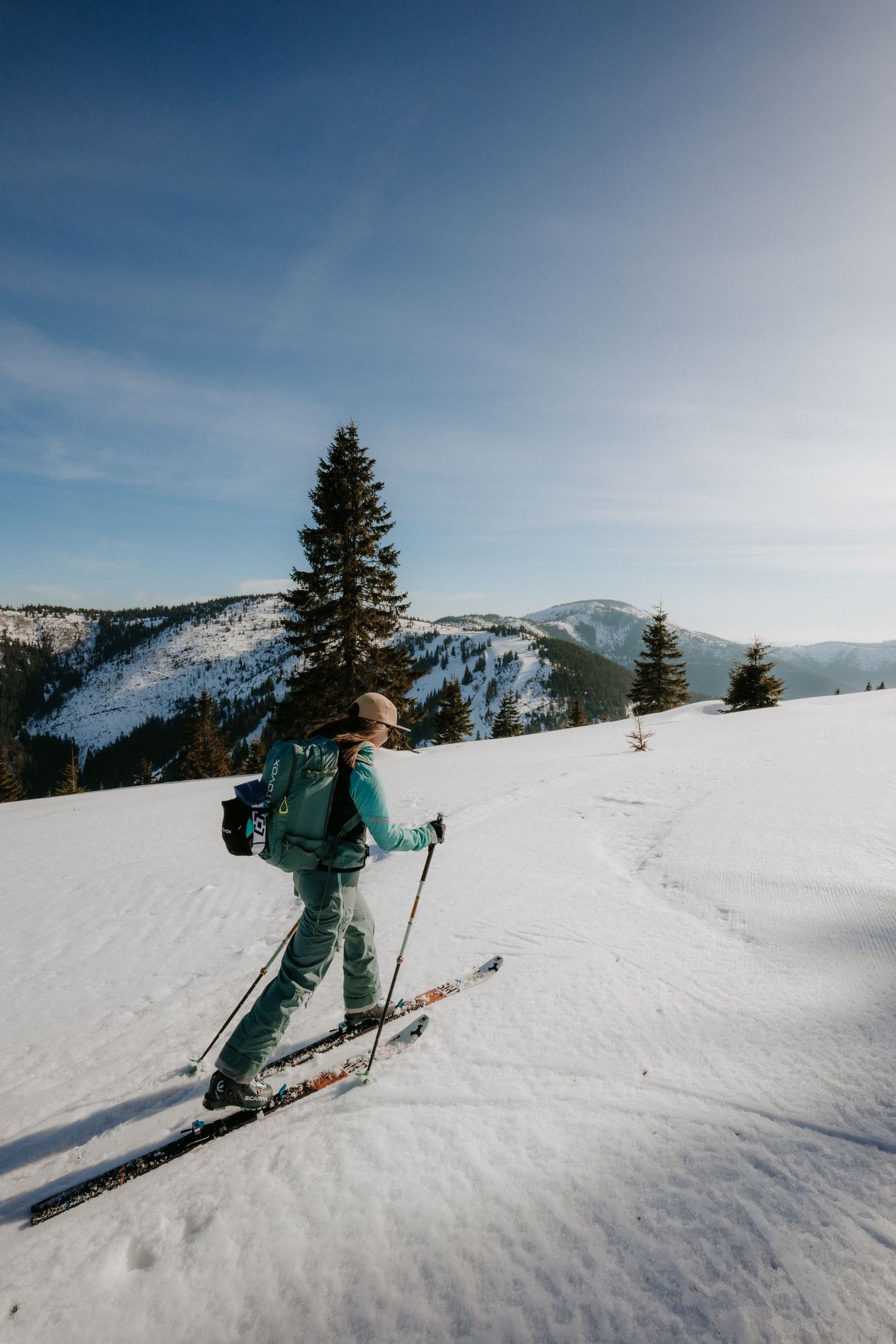 Woman in teal ski gear cross-country skiing on open snowy slope with mountain views.