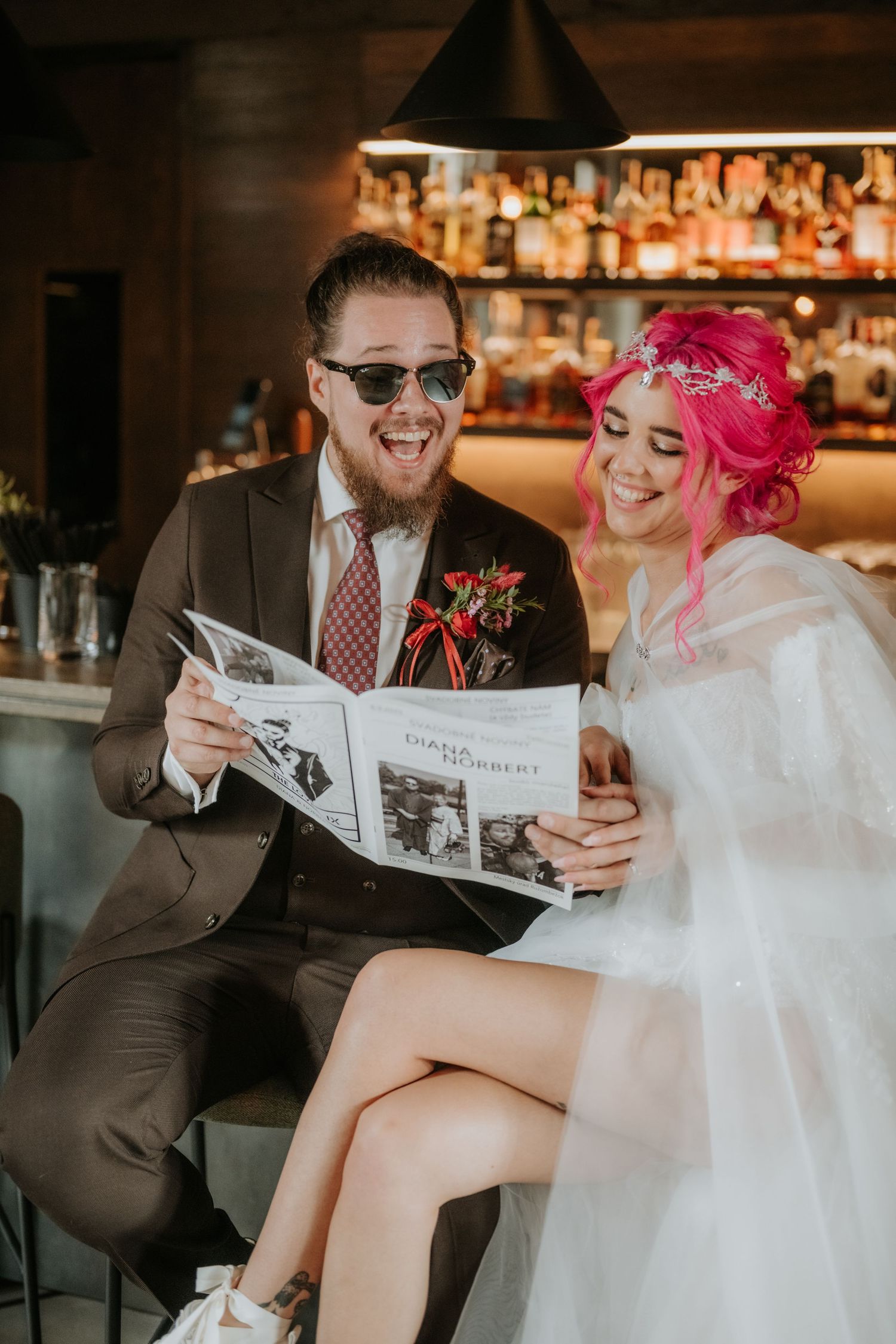 Laughing bride with pink hair and groom in sunglasses reading a magazine together at a bar on their wedding day.