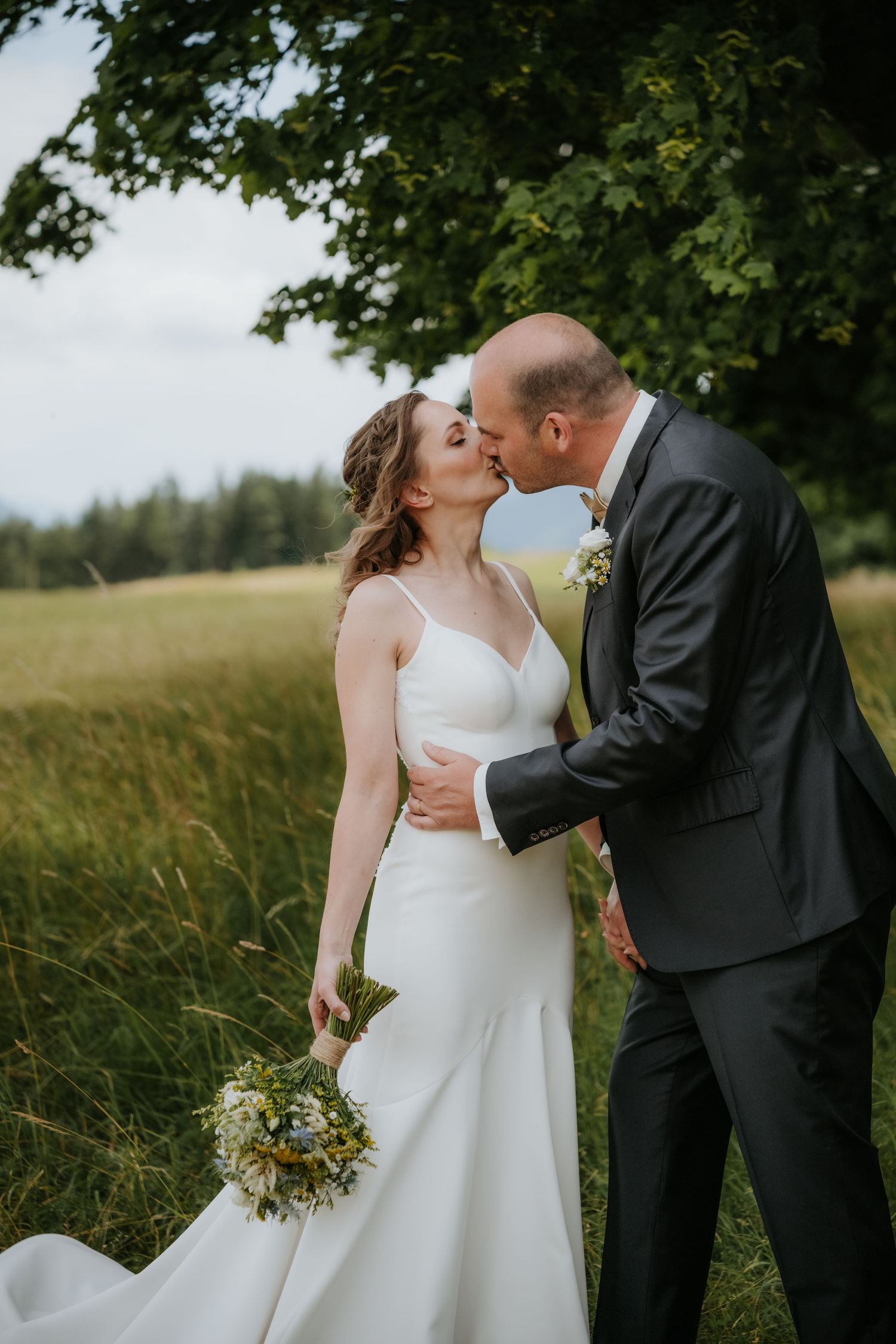 A bride and groom share a romantic kiss outdoors in a green meadow, she holds a wildflower bouquet in a white gown.