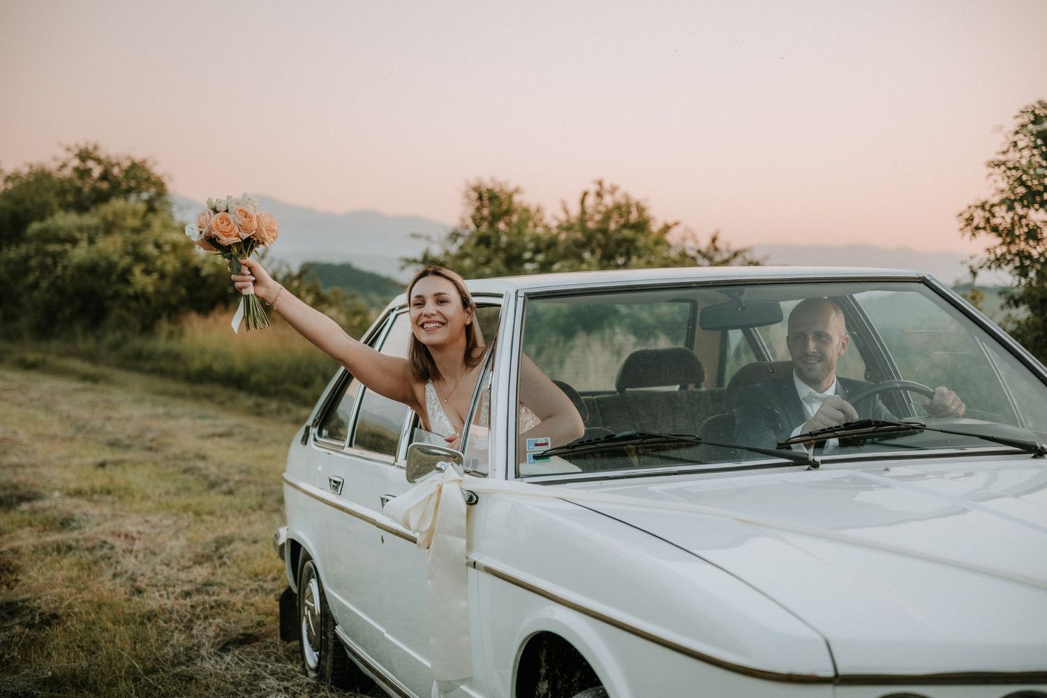 Bride leans out of white vintage car holding bouquet at sunset while groom drives through scenic countryside.