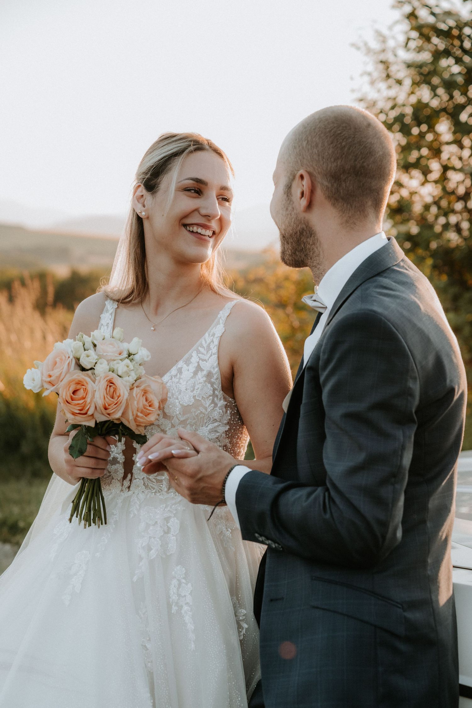 Smiling bride holds peach rose bouquet while groom gazes at her during outdoor golden hour wedding.
