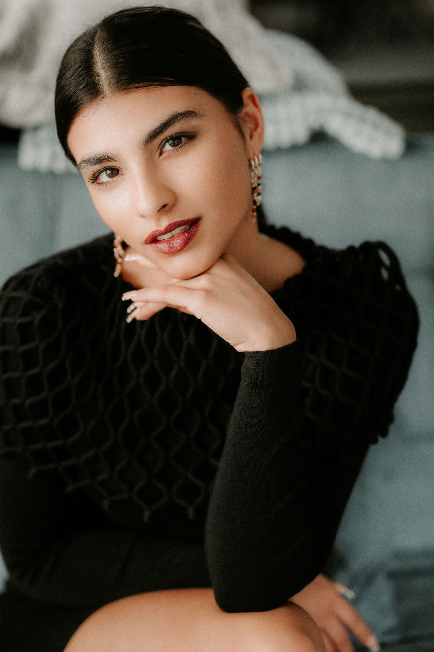 Young woman in a black textured sweater resting her chin on her hand, wearing statement earrings in a moody setting.