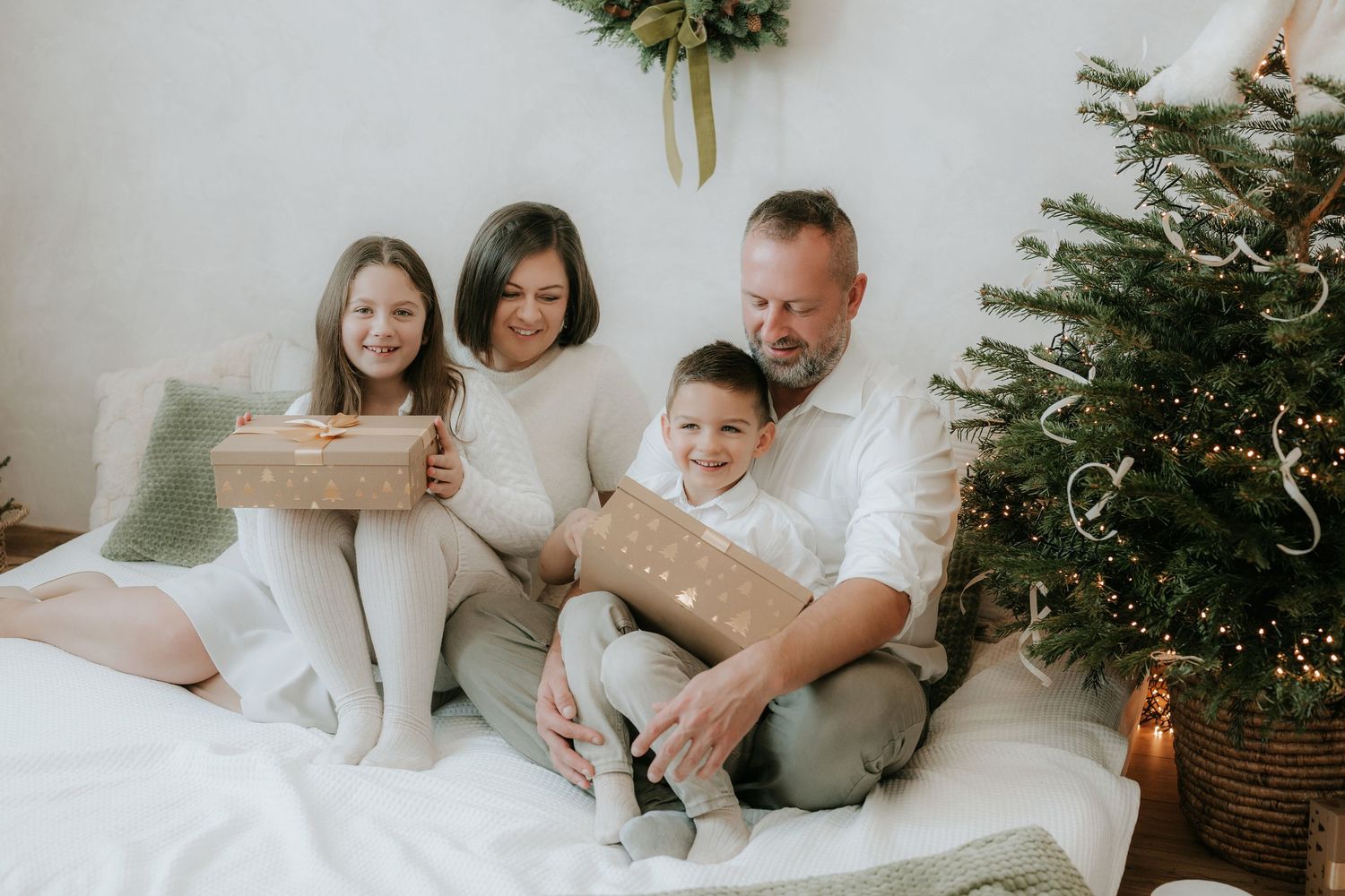 A happy family of four sits together near a Christmas tree, holding wrapped gifts in a festive holiday setting.