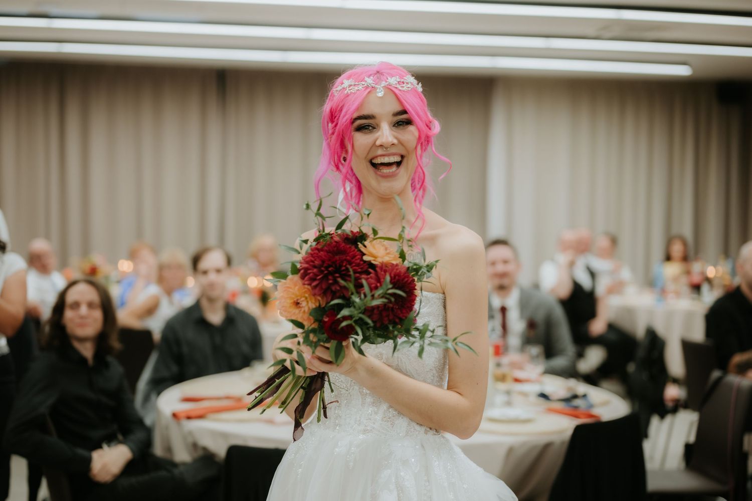 Joyful bride with pink hair holding a colorful bouquet laughs at her wedding reception in a banquet hall.
