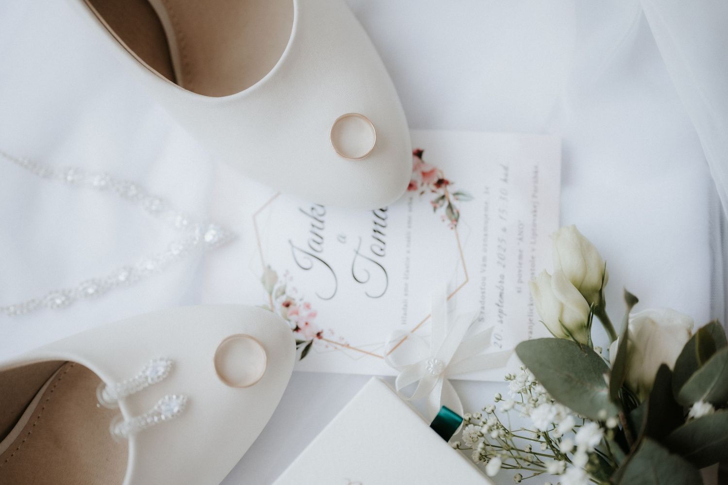 Elegant wedding flat lay with gold rings, pearl necklace, bridal shoes, floral invitation, and white roses on white fabric.