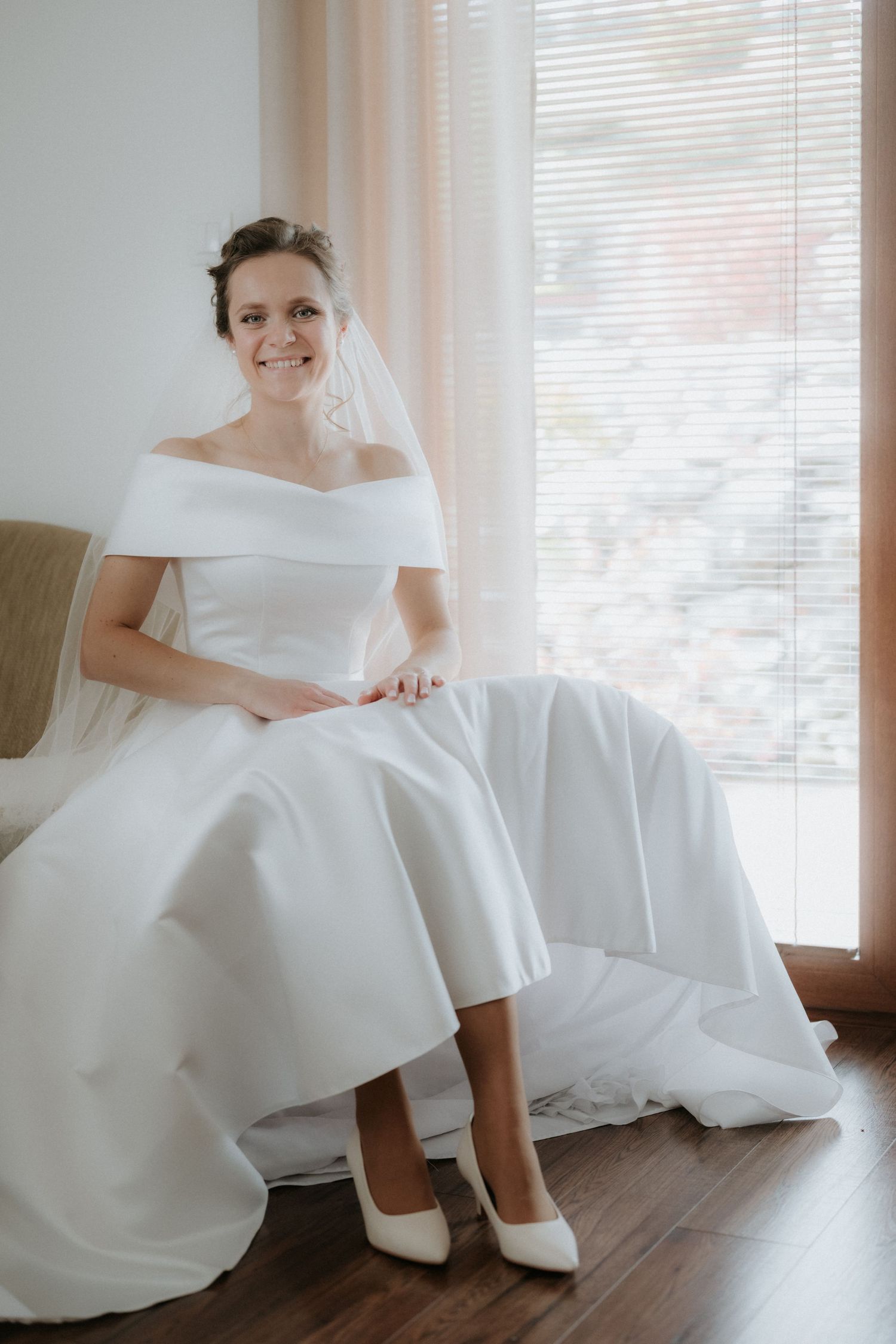 Smiling bride in off-shoulder white gown and veil sits gracefully by a bright window in white heels.