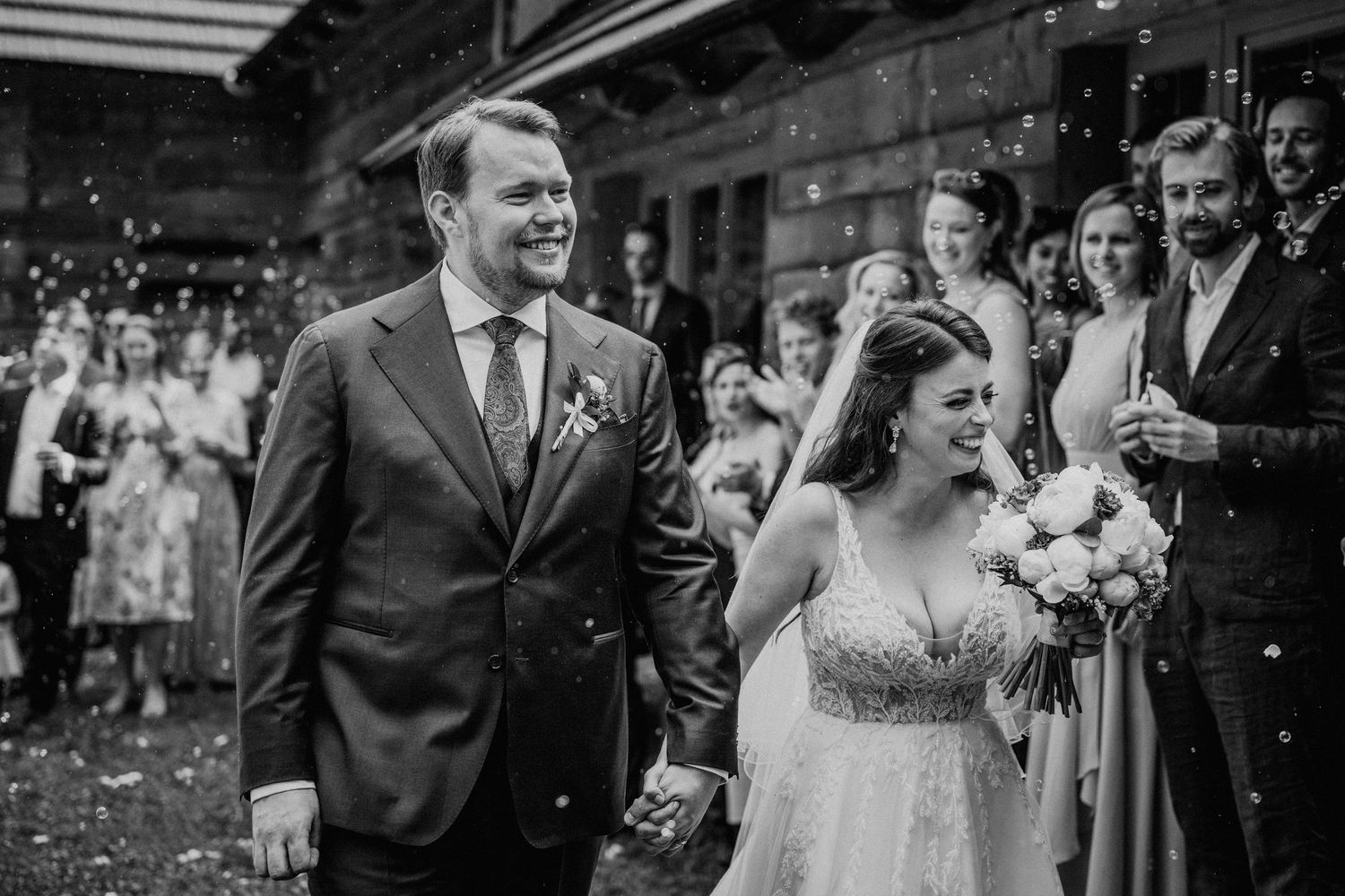 Black and white photo of a smiling bride and groom walking through a bubble send-off after their wedding ceremony.