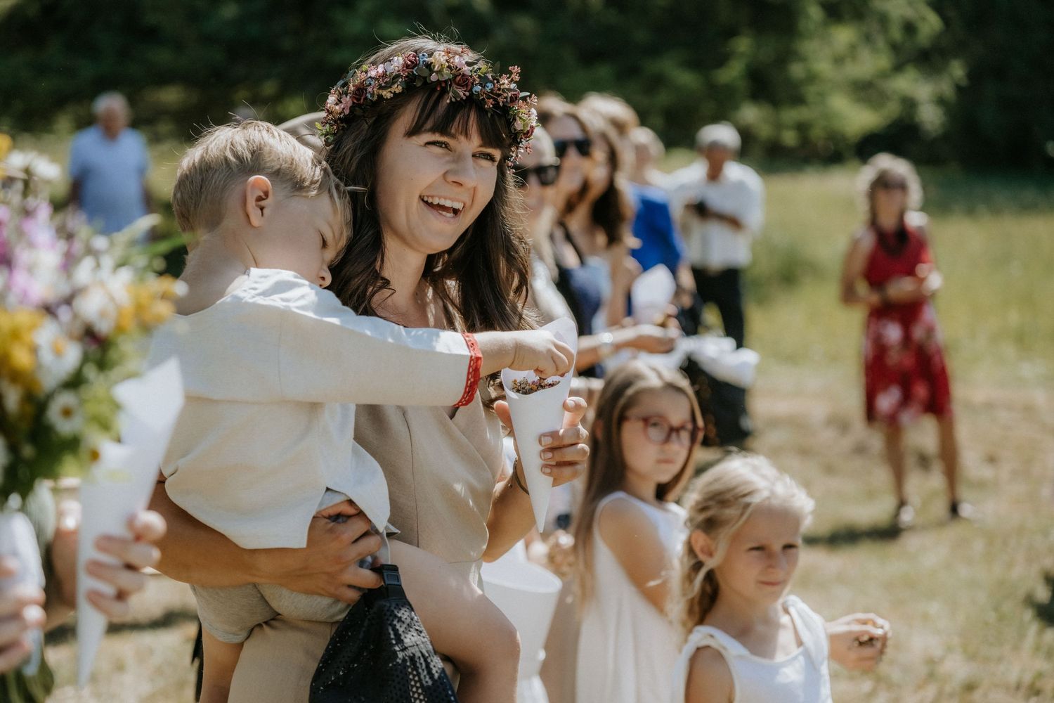 Woman with flower crown holding baby laughs joyfully at outdoor wedding ceremony with children and guests.