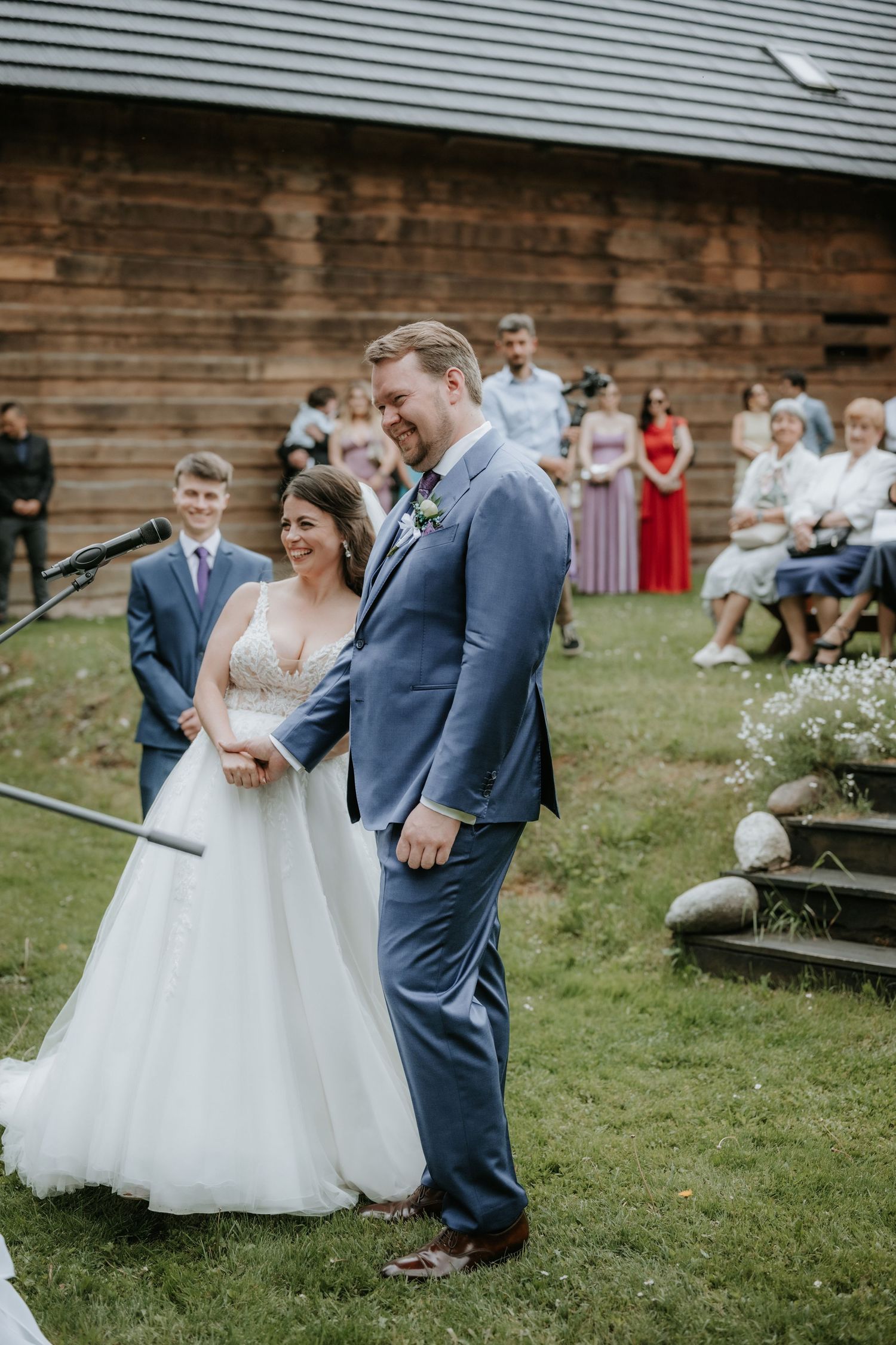Bride in white gown and groom in blue suit laughing during outdoor wedding ceremony near rustic wooden barn.