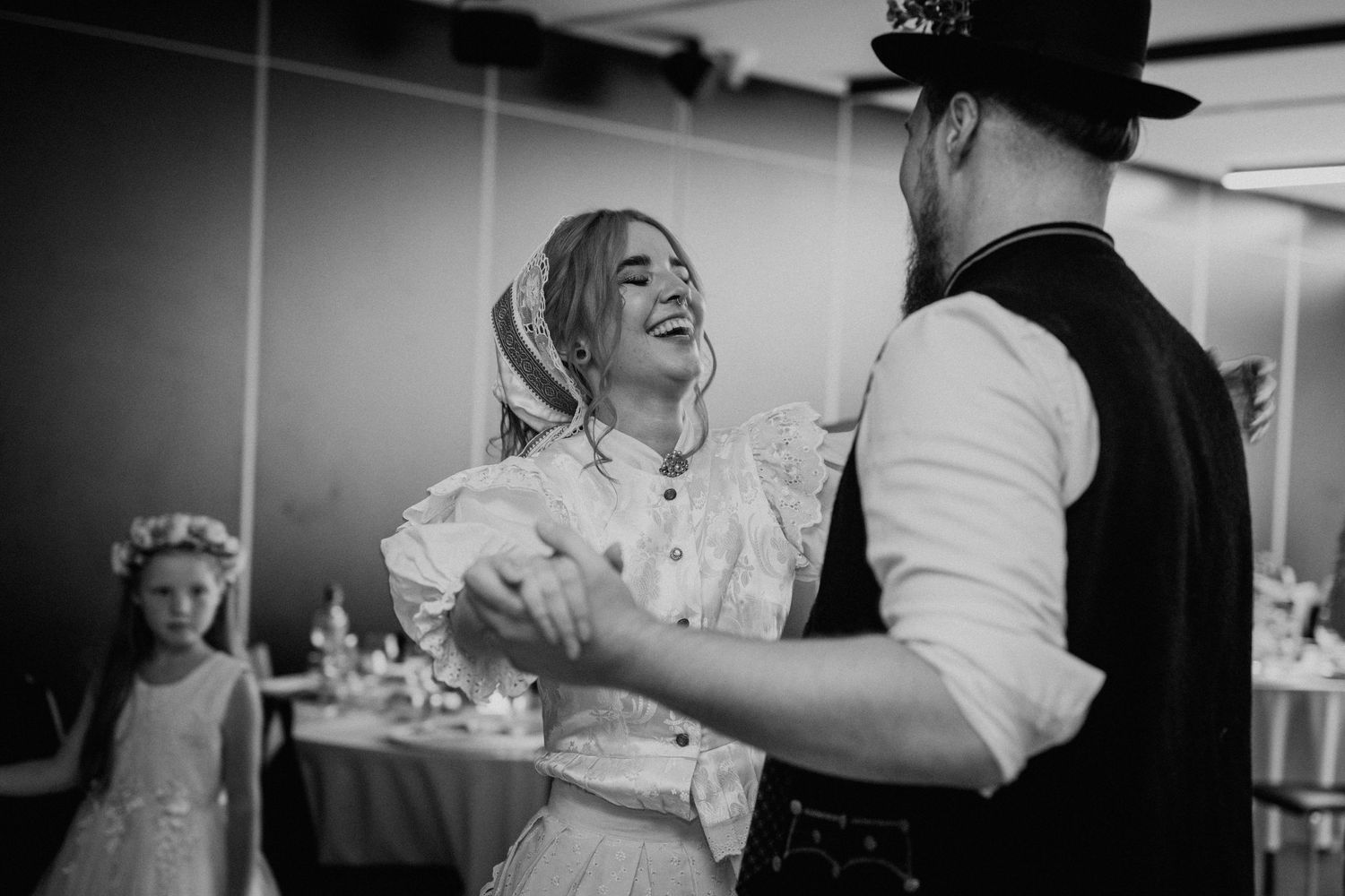 Black and white photo of a couple dancing joyfully at a wedding reception, with a flower girl in the background.