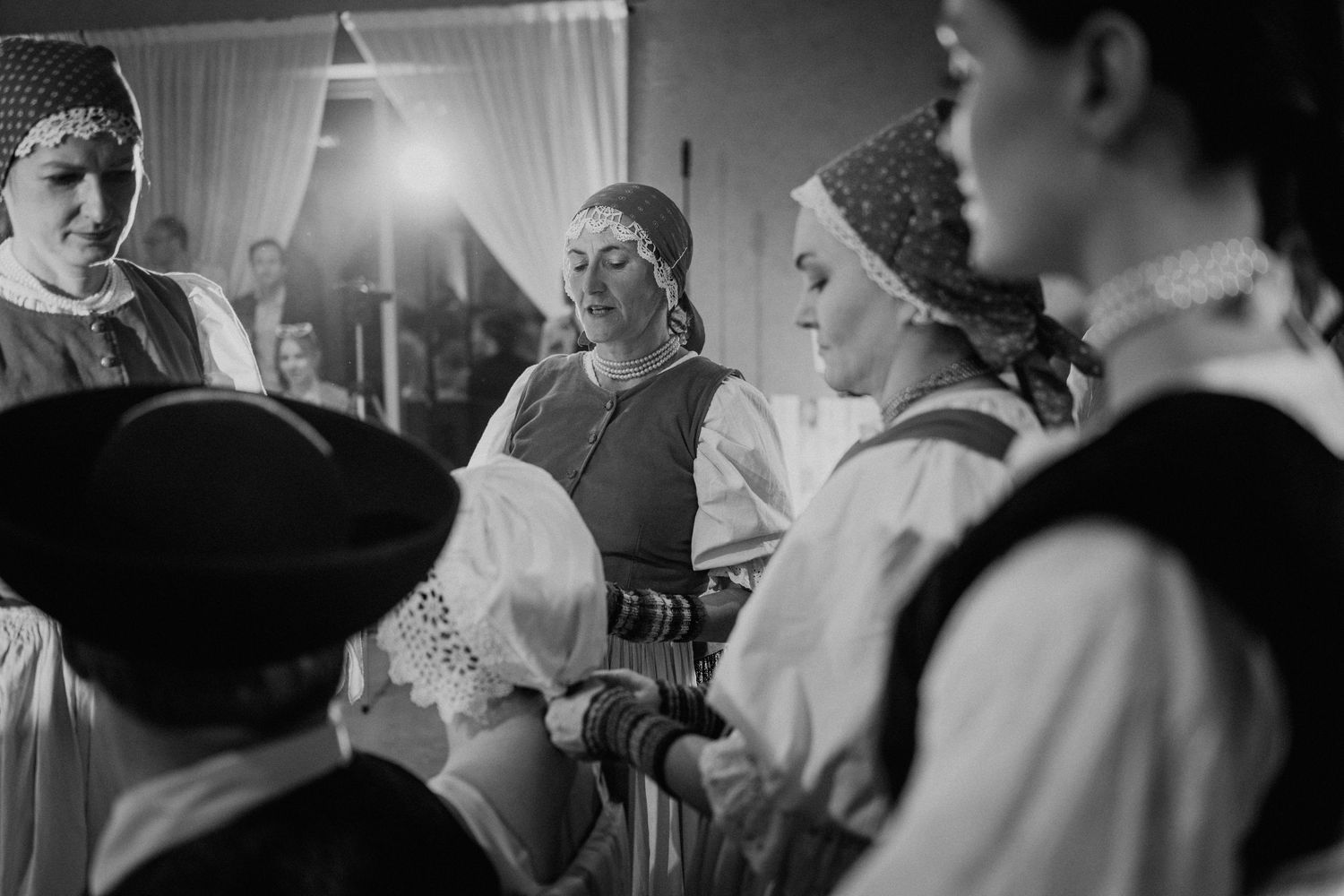 Black and white photo of women in traditional folk costumes performing a cultural dance ceremony indoors.