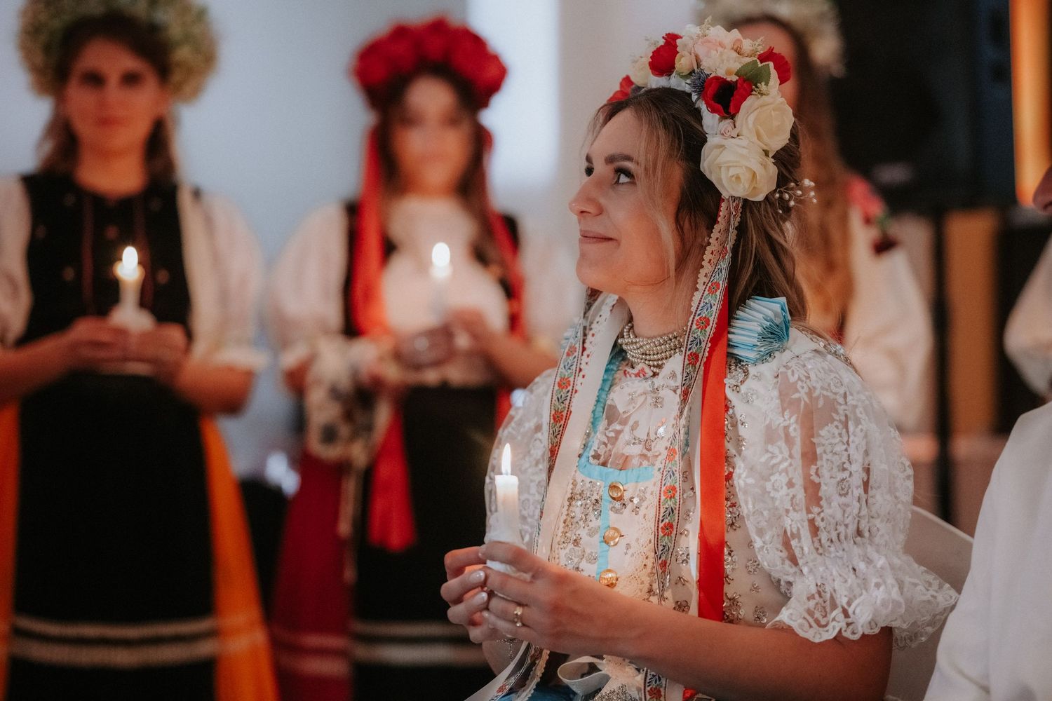 Woman in traditional Ukrainian embroidered dress holding candle at cultural ceremony with floral crown.